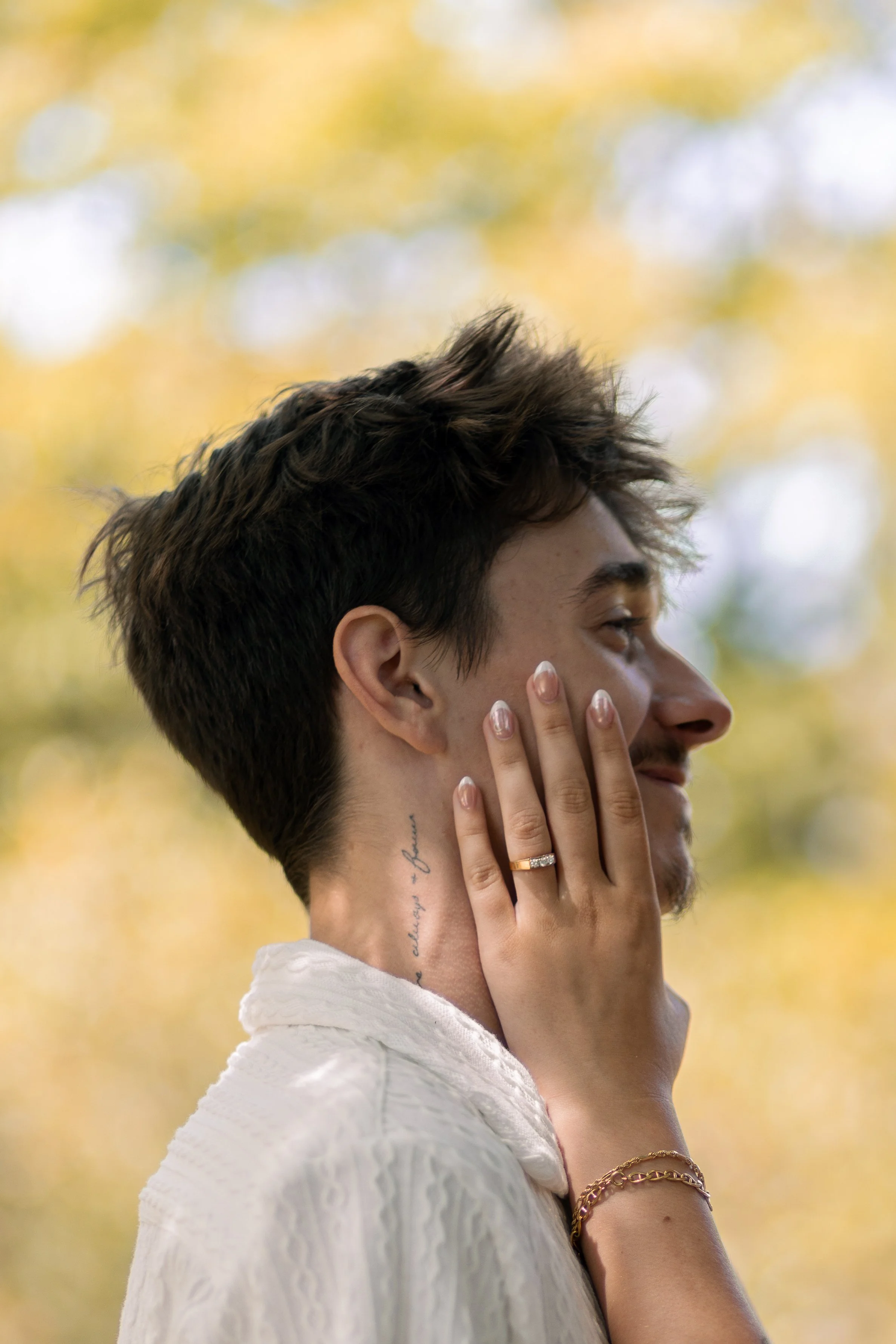A young man with dark, tousled hair and a tattoo on his neck, wearing a white textured shirt, is gently holding his face with a woman's hand, which has a wedding ring. The woman has a gold bracelet on her wrist. The background shows blurred autumn le