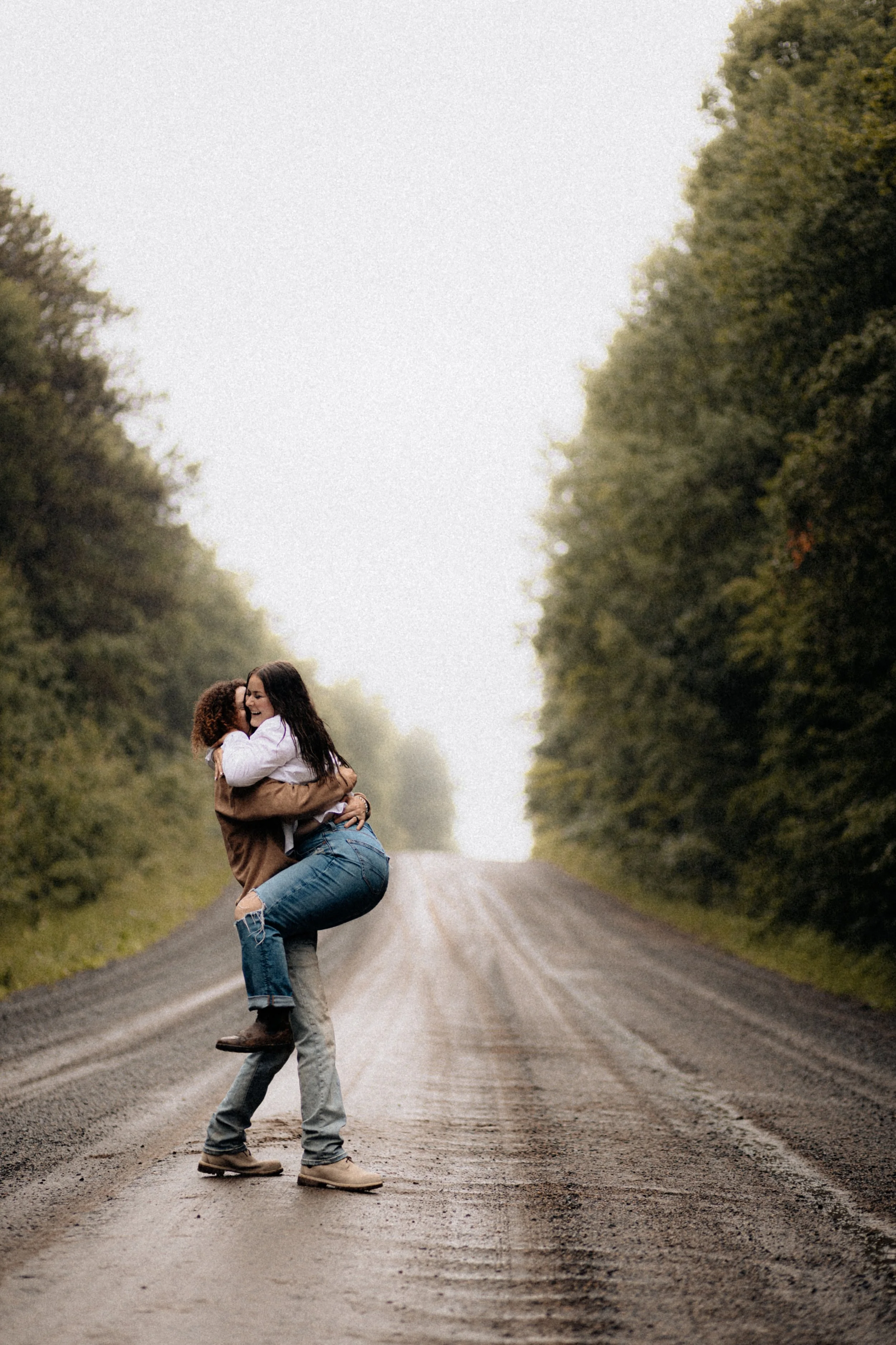 Two women embracing and smiling on a rural dirt road surrounded by trees.