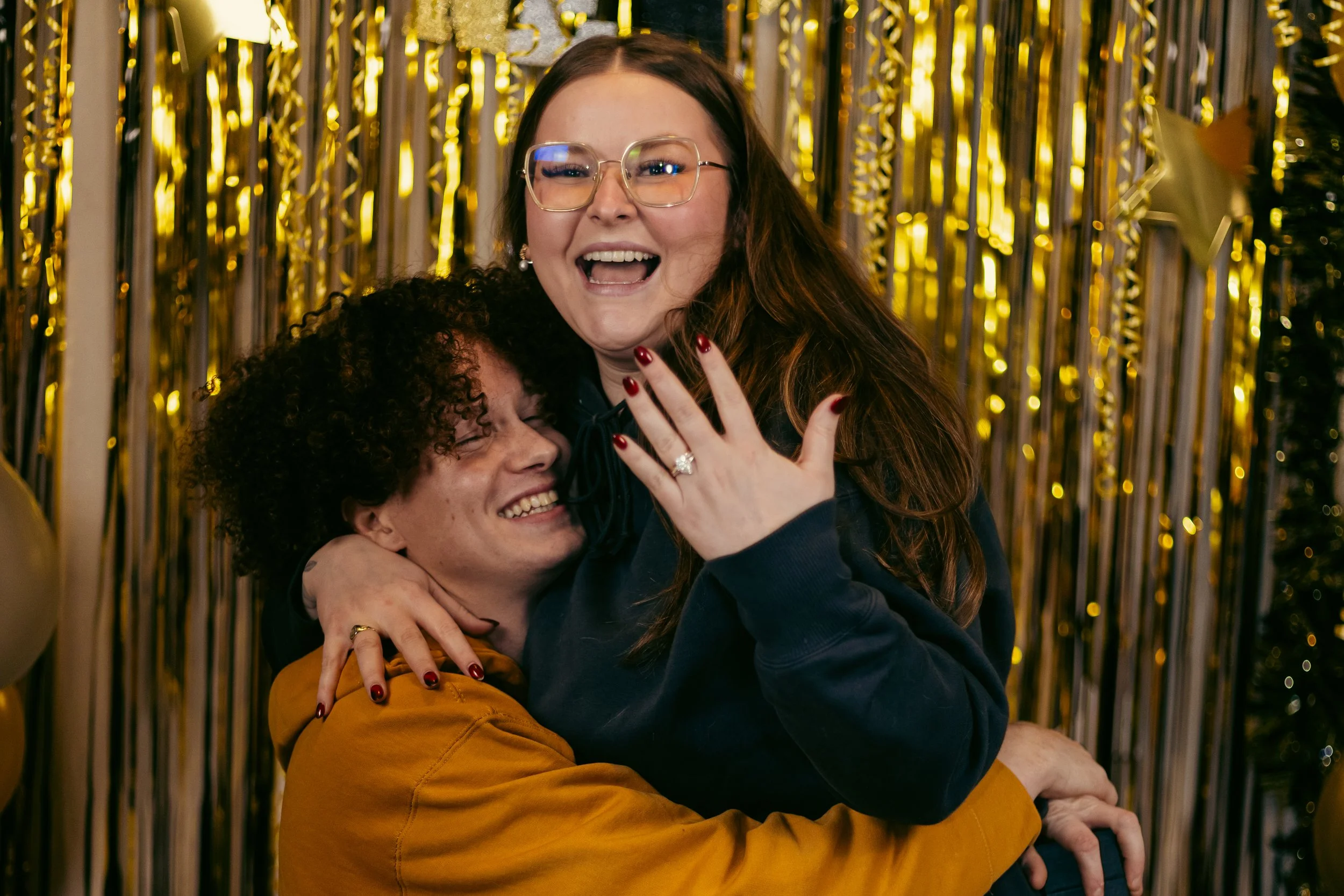 A joyful couple hugging and celebrating, with a woman showing her engagement ring, in front of gold and silver decorations.