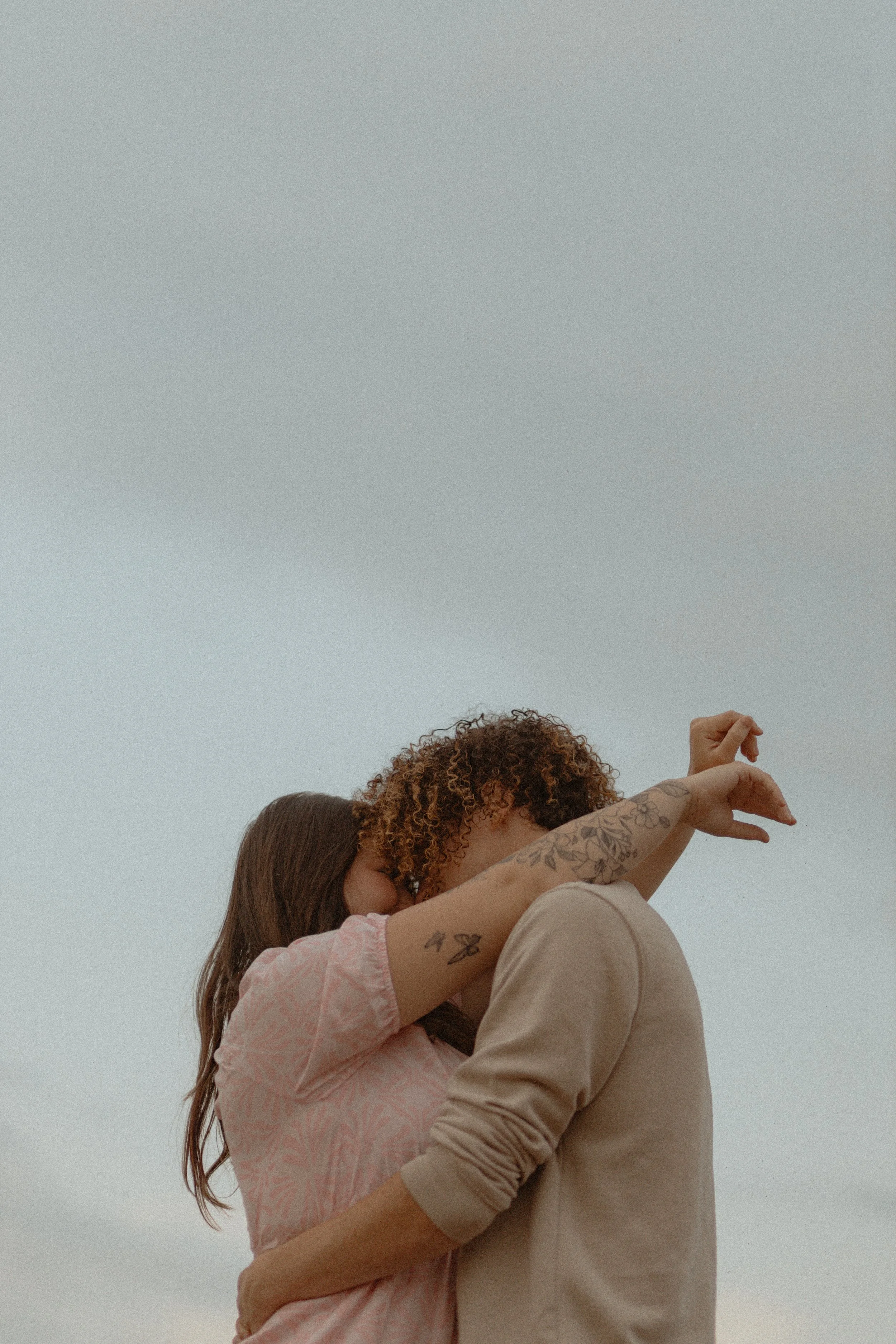 A couple embracing outdoors, with their foreheads touching and arms wrapped around each other. The woman has tattoos on her arm and long brown hair, and the man has curly hair. The background features an overcast sky.