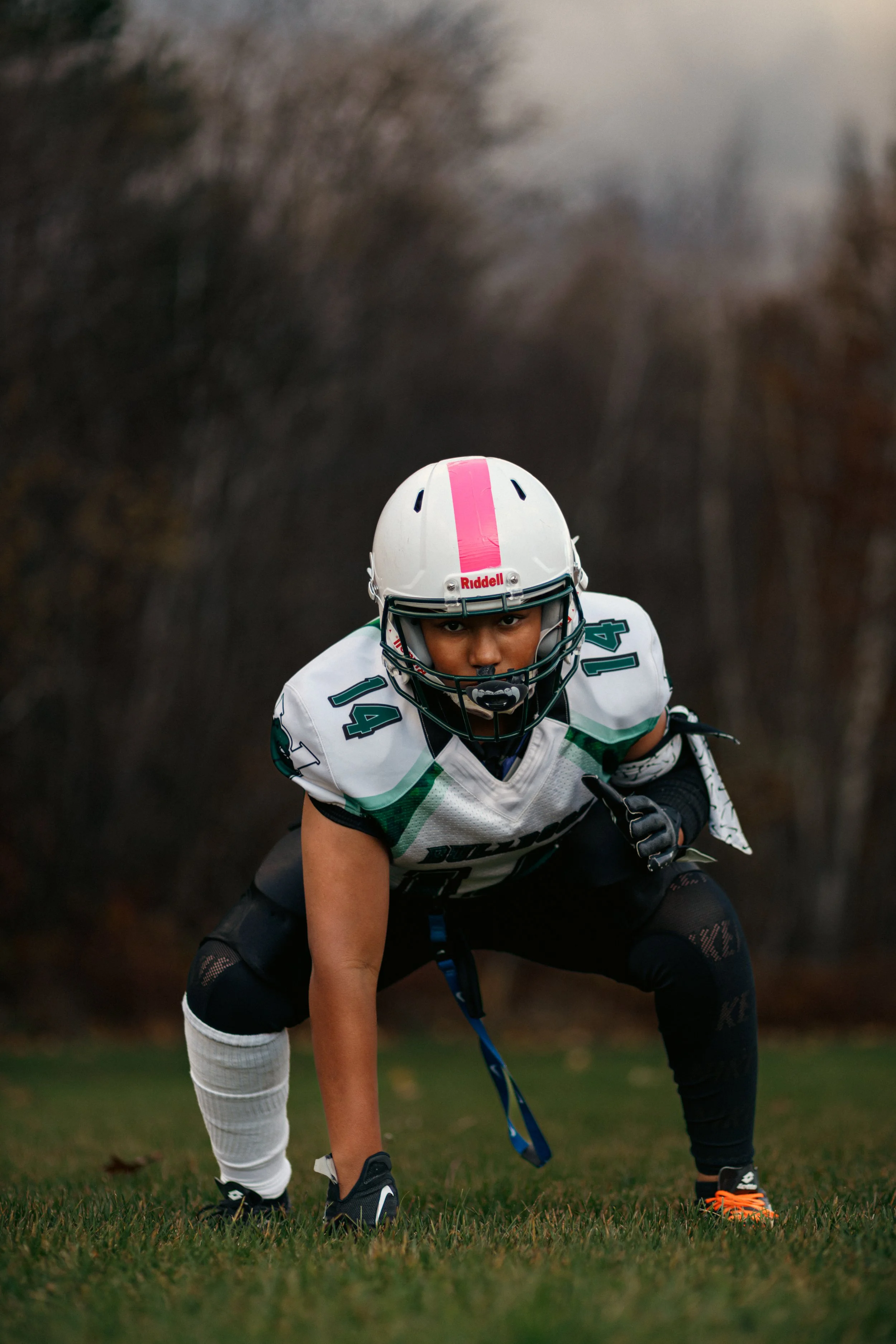 A football player in a white jersey with green and black accents, wearing a white helmet with pink stripe, crouched in a ready stance on a grassy field.