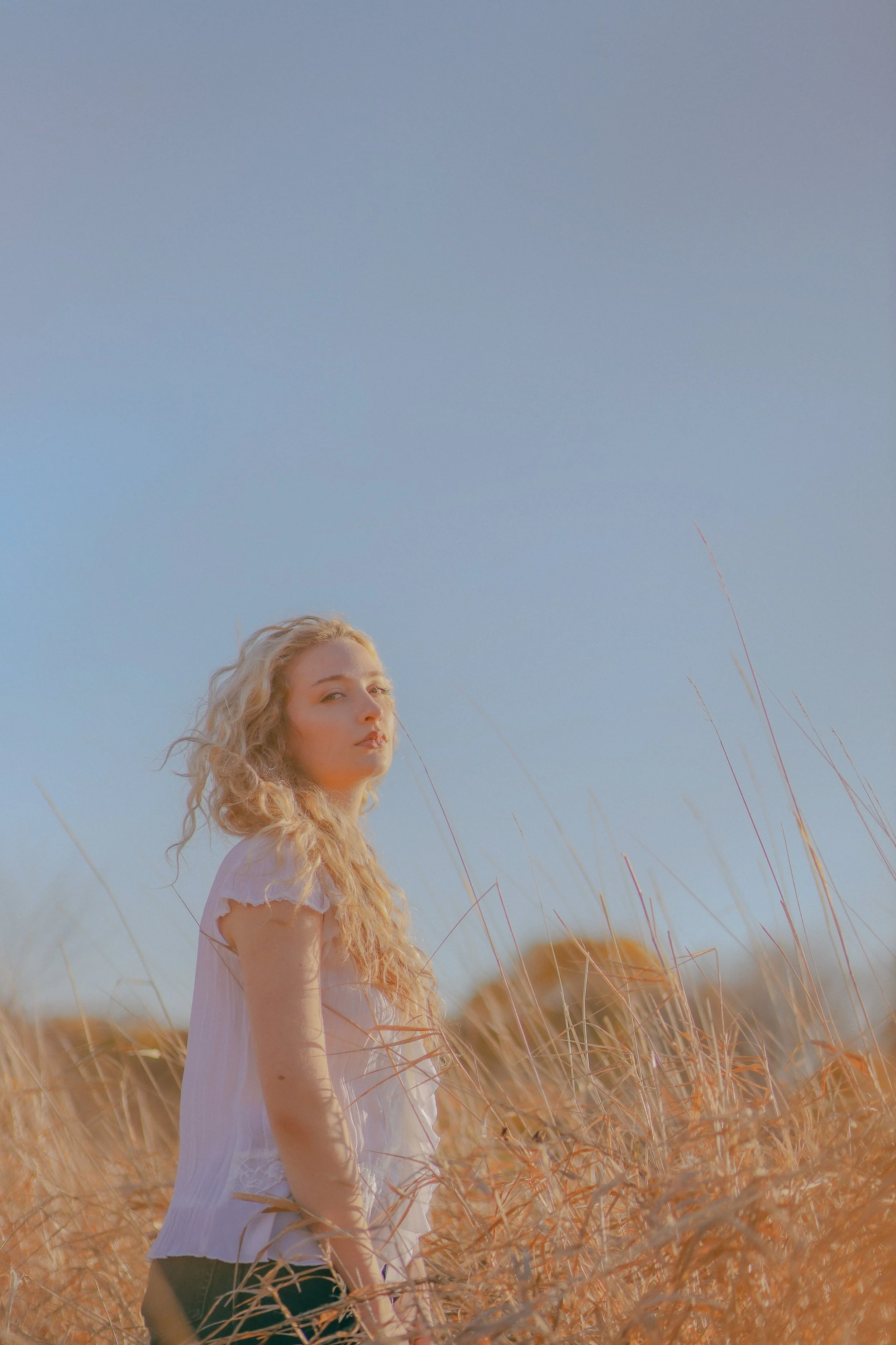 A woman with long curly blonde hair standing in a field of tall, golden grass under a clear blue sky.