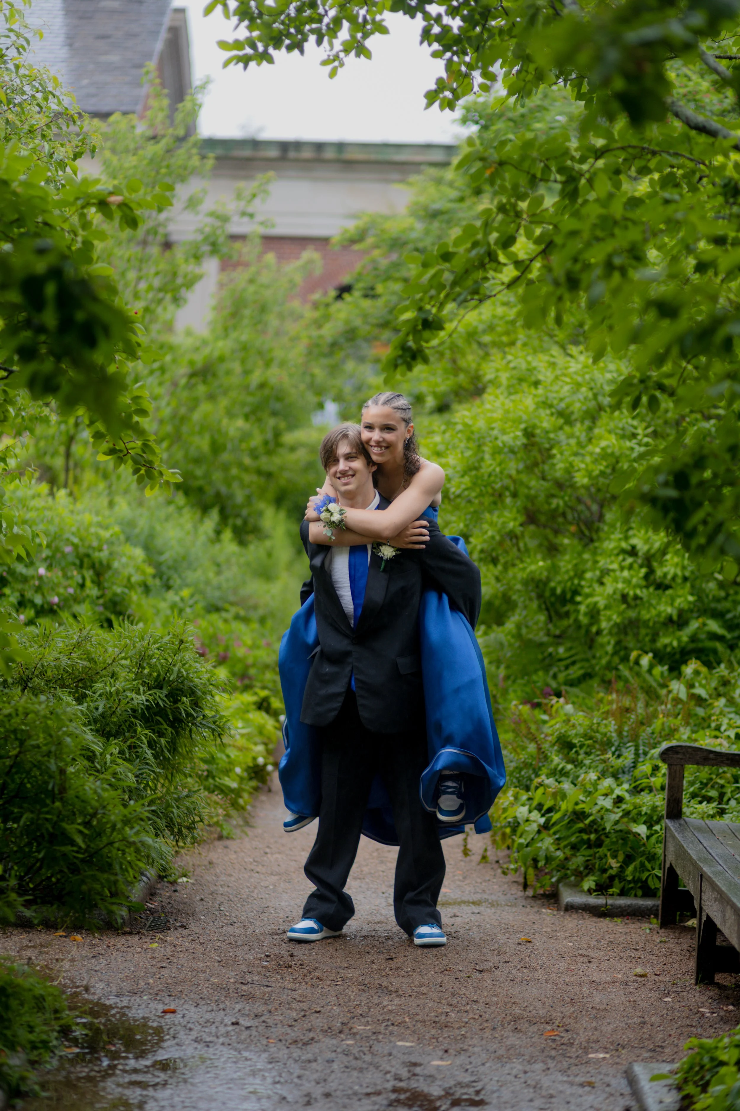 A young couple in formal attire, the woman in a blue gown and the man in a black suit, sharing a playful piggyback ride on a garden pathway surrounded by lush green trees and bushes.