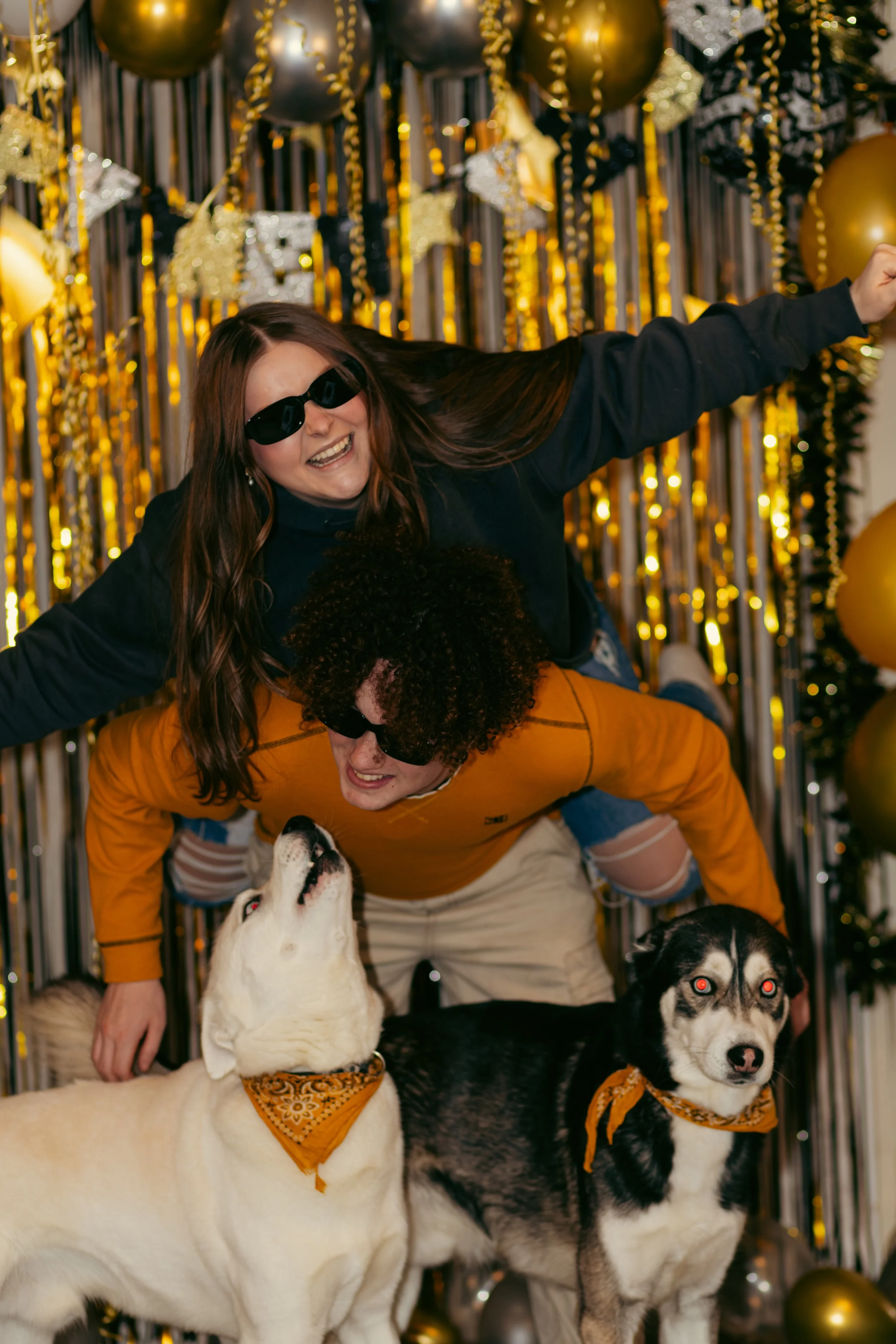 People dressed in casual clothes and sunglasses, with two dogs, celebrating at a party with gold and black balloons and streamers.