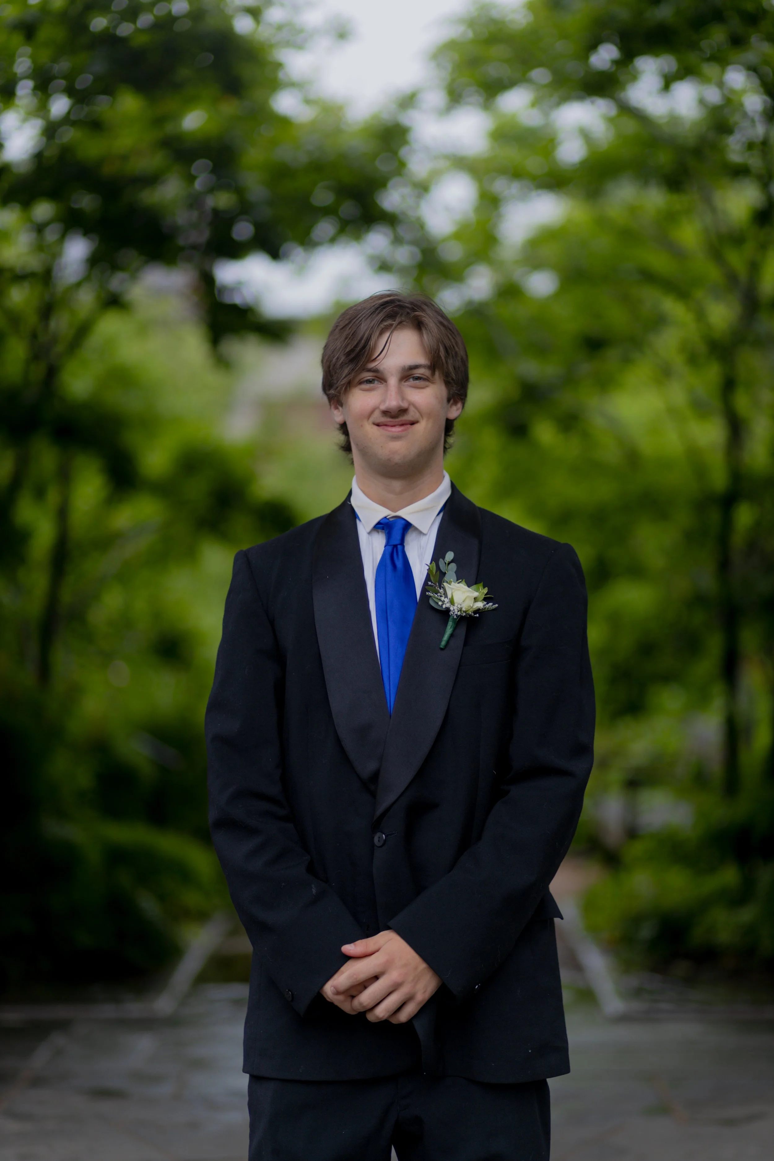 Young man in formal tuxedo with blue tie and boutonnière, standing outdoors in front of green trees.