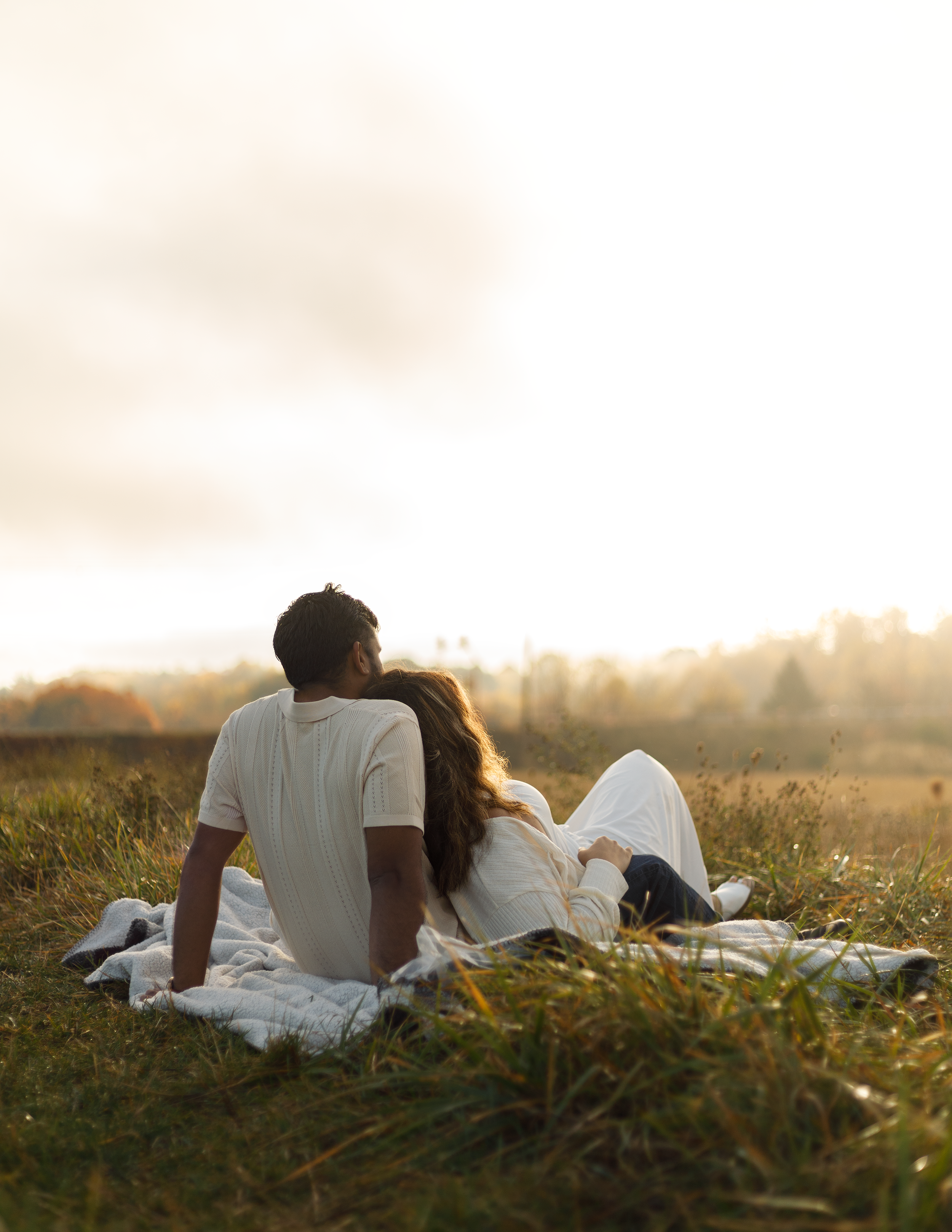 A couple sitting on a blanket in a grassy field during sunset, with trees and a cloudy sky in the background.