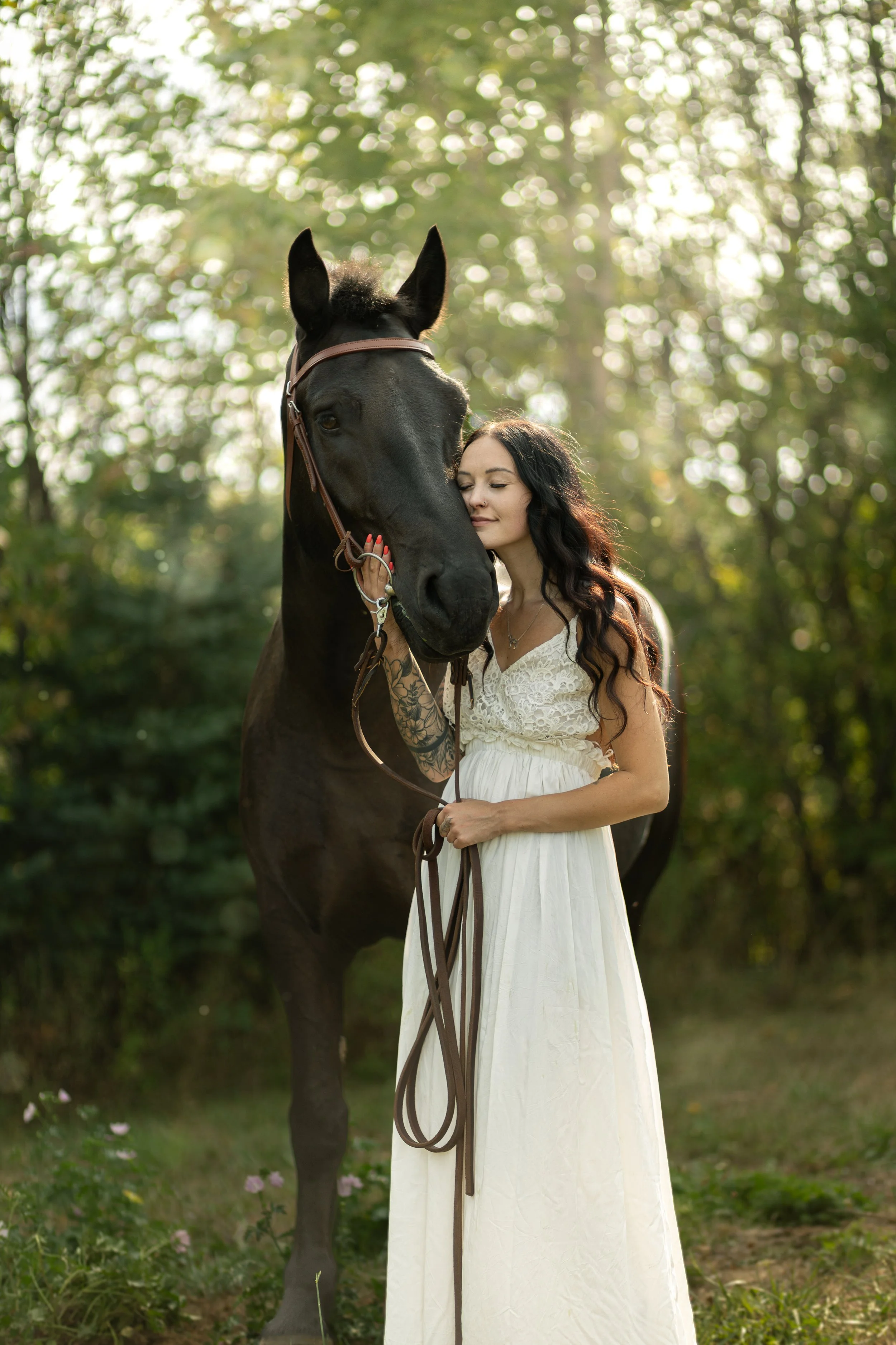 A woman with dark wavy hair and a white dress gently leans her head against the nose of a large black horse outdoors in a natural setting with trees and sunlight.