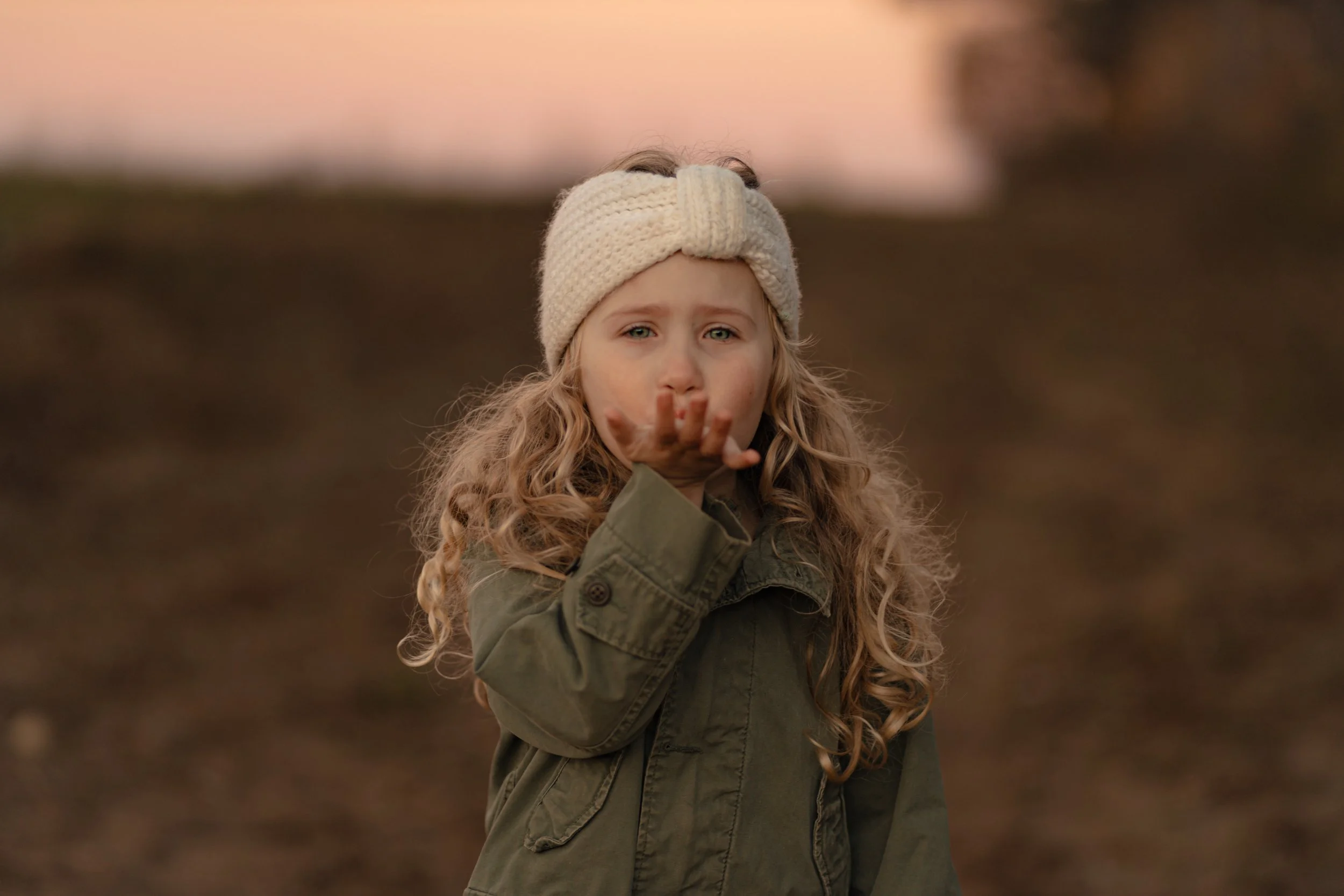 A young girl with curly blonde hair, standing outdoors in a natural setting during sunset, wearing a beige knit headband and an olive green jacket, with her hand extended towards the camera as if blowing a kiss.