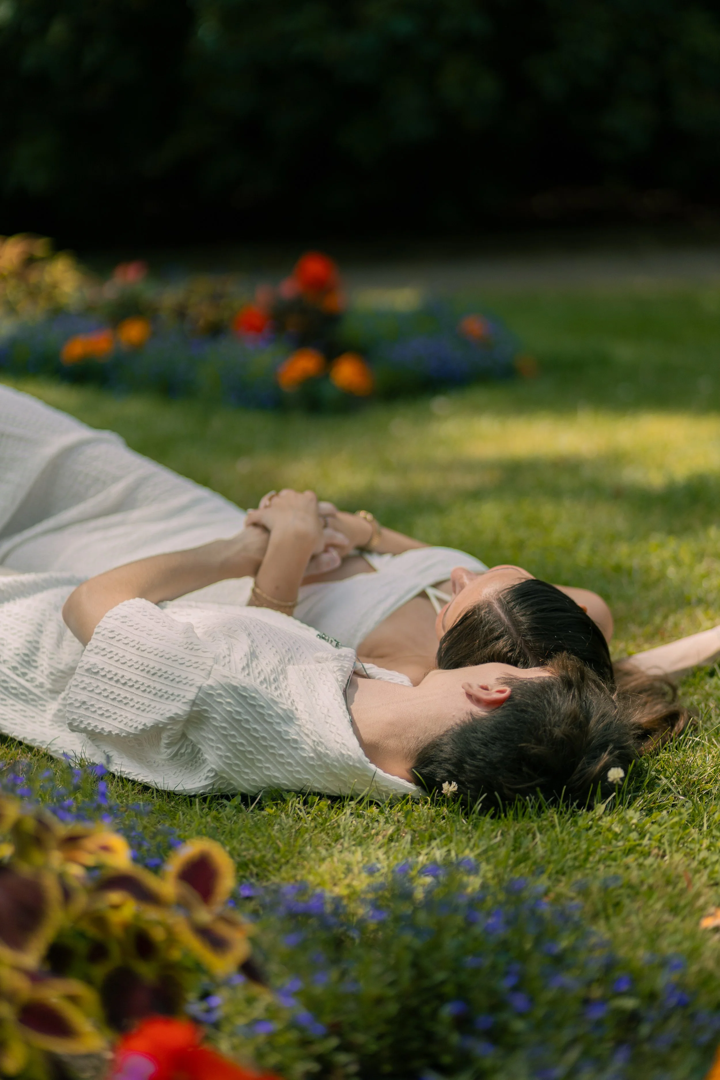 A couple lying on grass in a park with flowers and trees in the background.