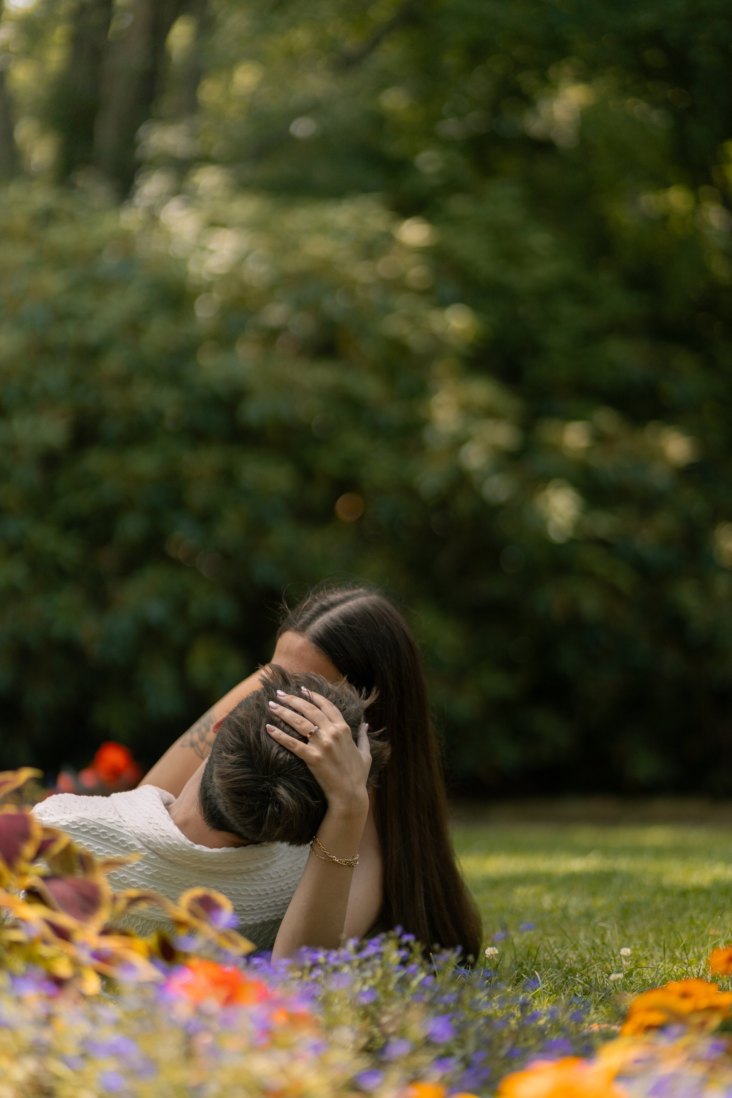 A woman and a boy lying on the grass in a park with flowers in the foreground, embracing each other.