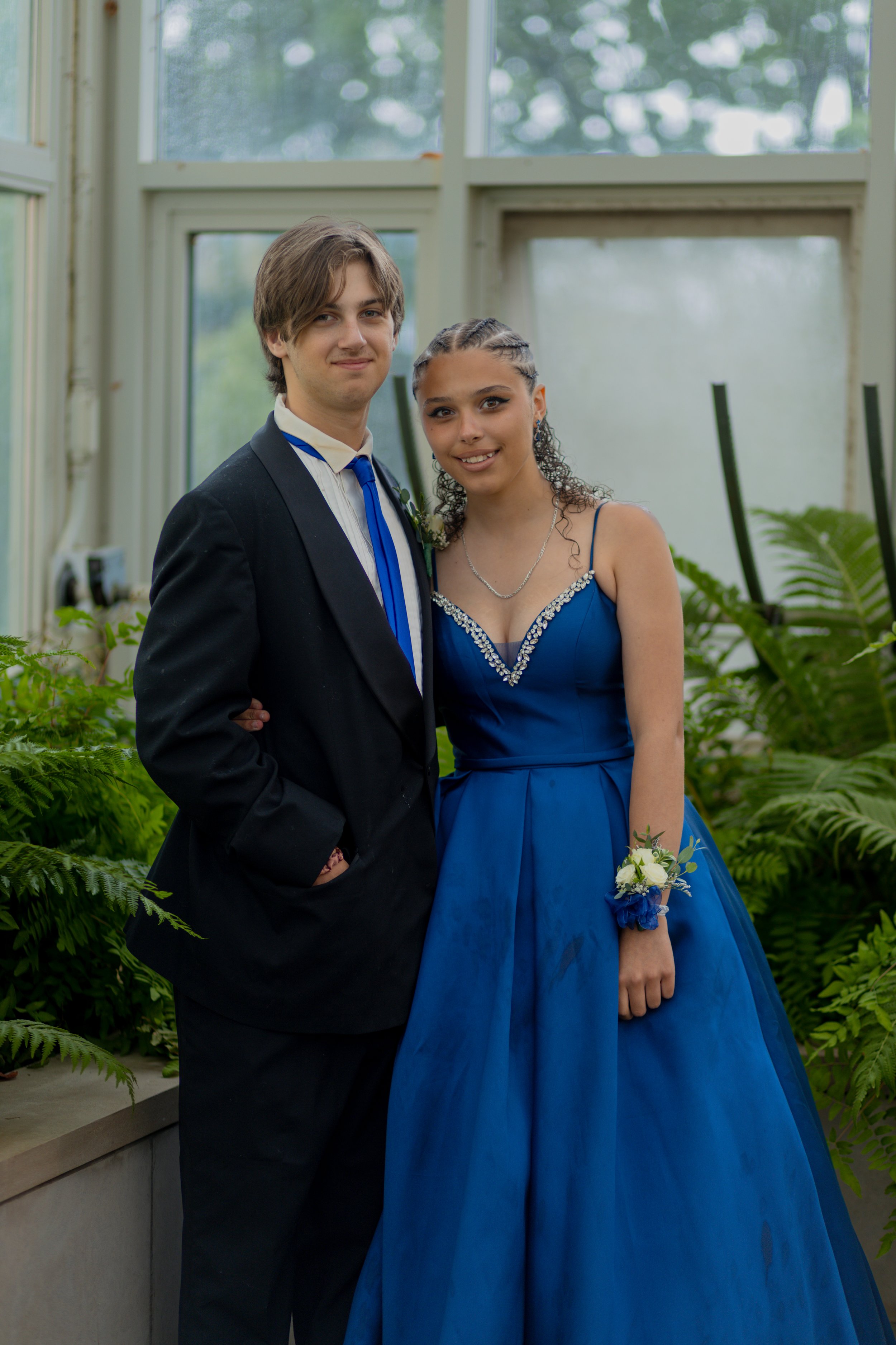 Young man in black suit and young woman in blue gown standing together indoors with lush green plants and large windows in the background.
