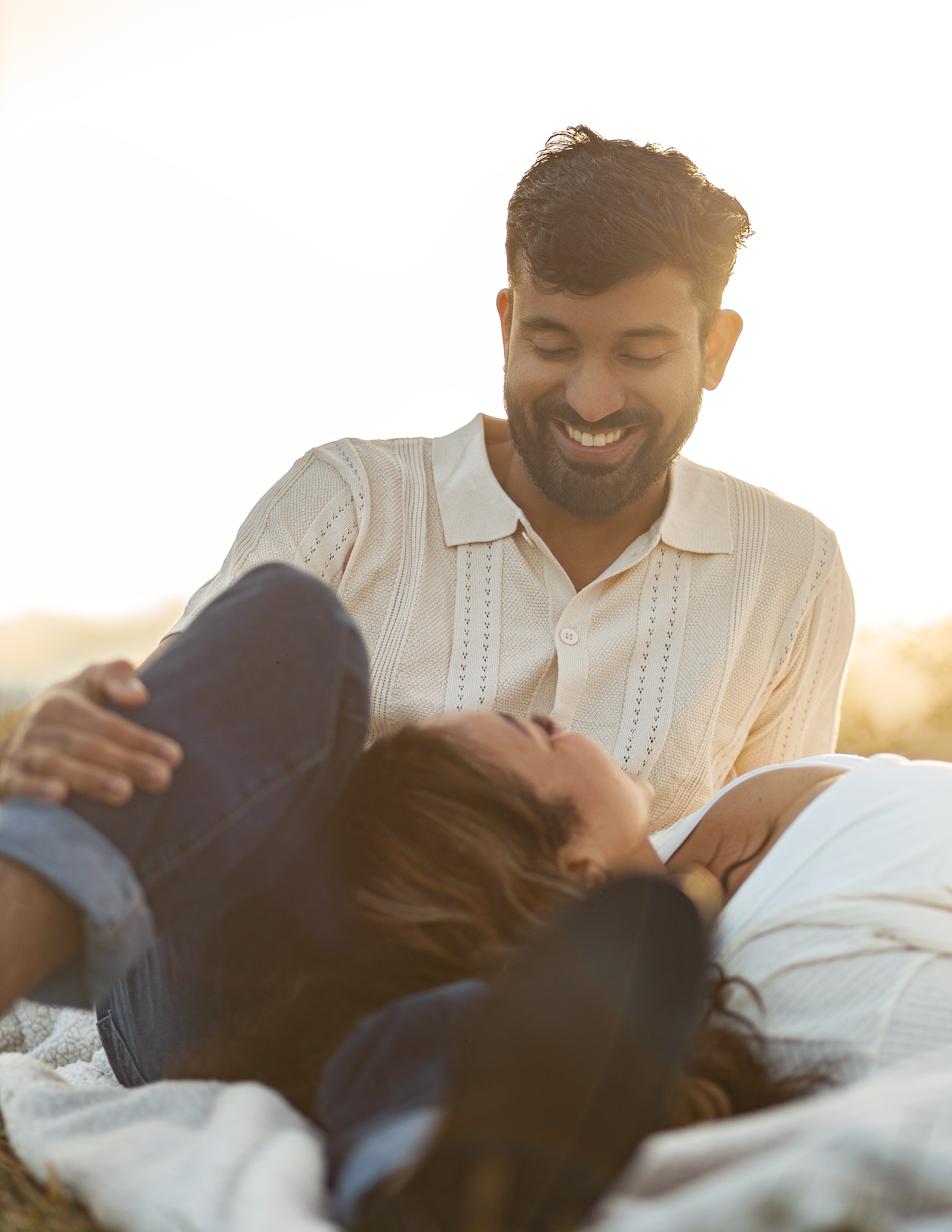 A man and woman enjoying a moment outdoors, with the man smiling at the woman who is lying on her back, wearing a hat and denim jacket, during sunset.