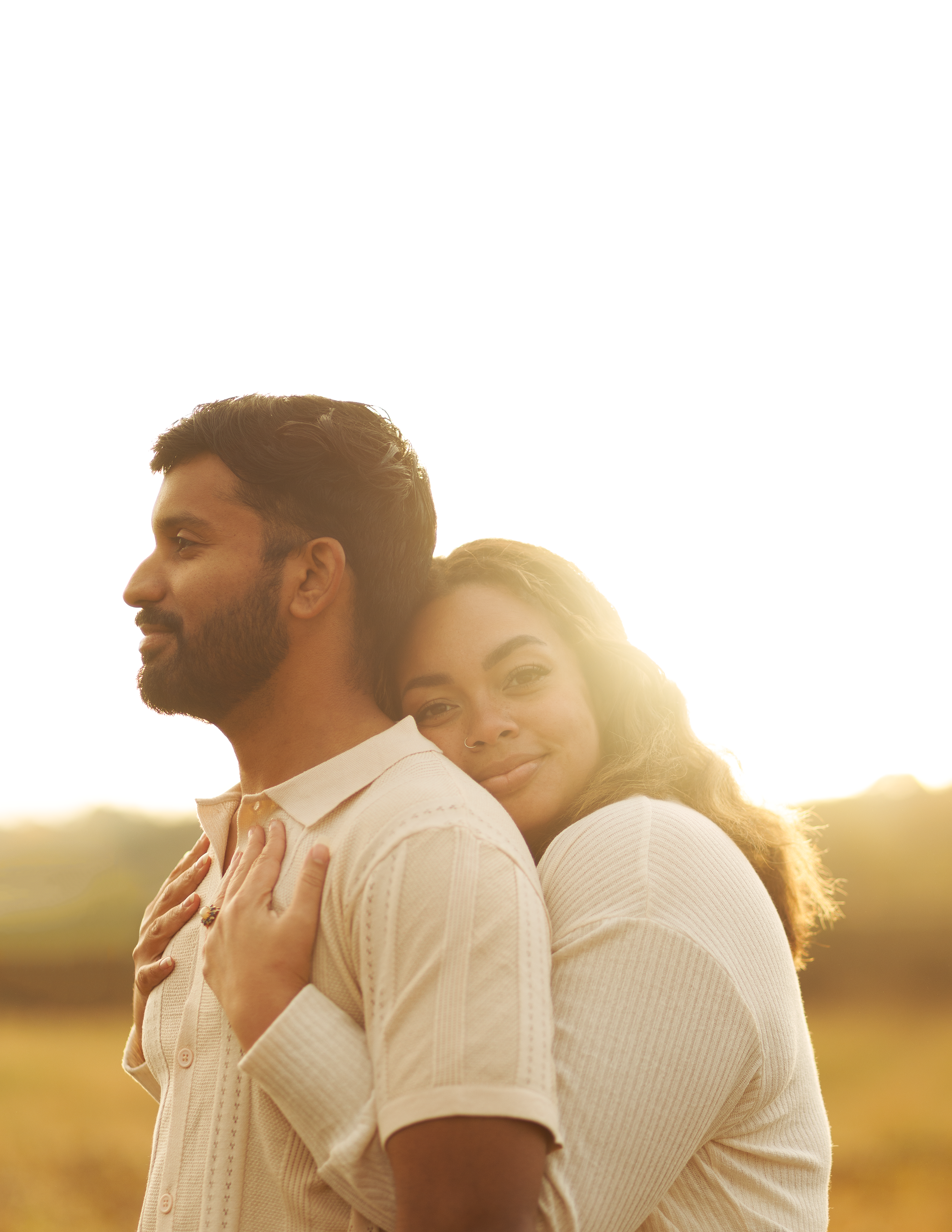 A couple standing close together outdoors at sunset, with the woman hugging the man from behind and smiling at the camera.