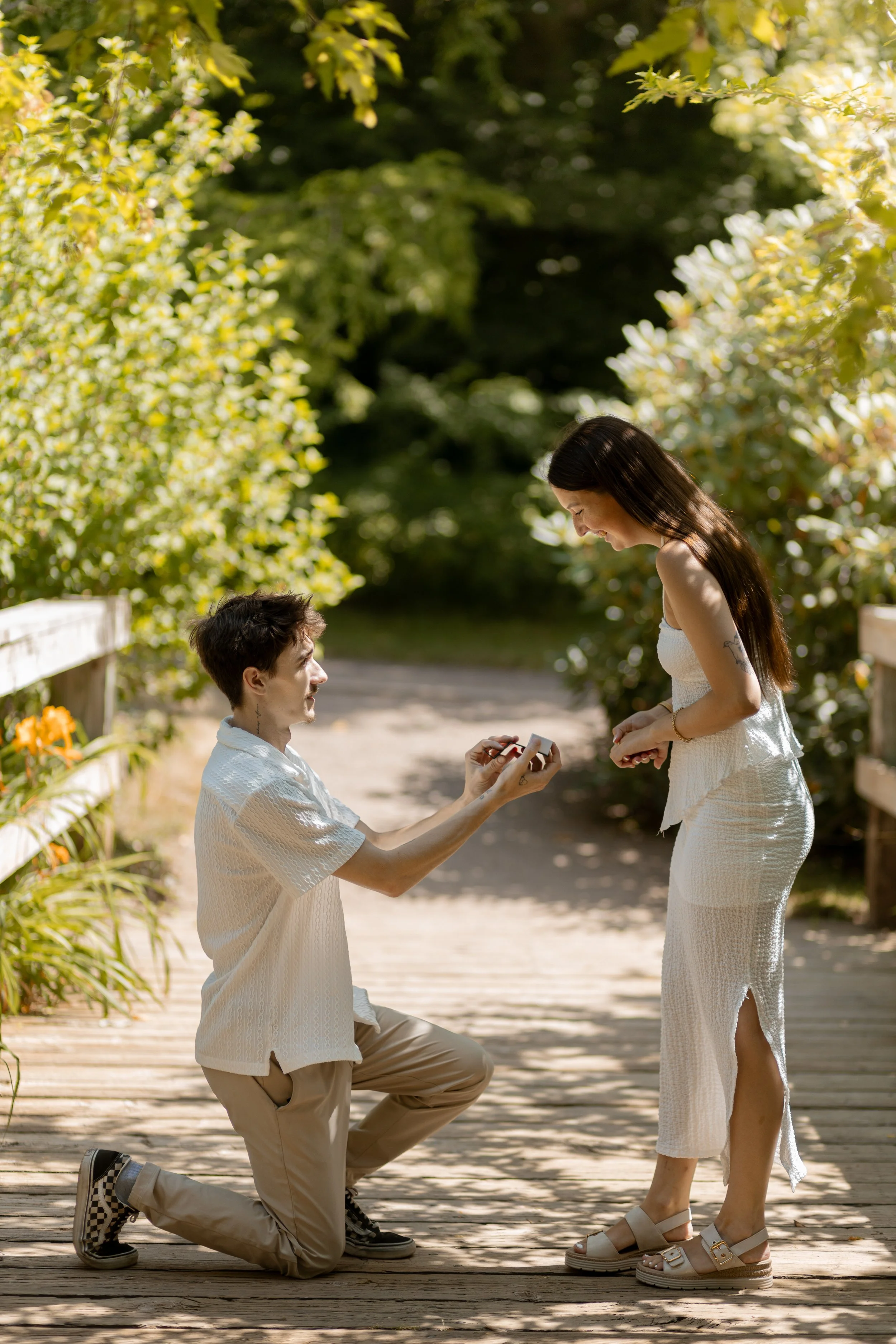 A man proposing marriage to a woman on a wooden pathway in a lush, green outdoor setting, both dressed in white, with the man kneeling and holding a ring box, the woman smiling and looking at the man.