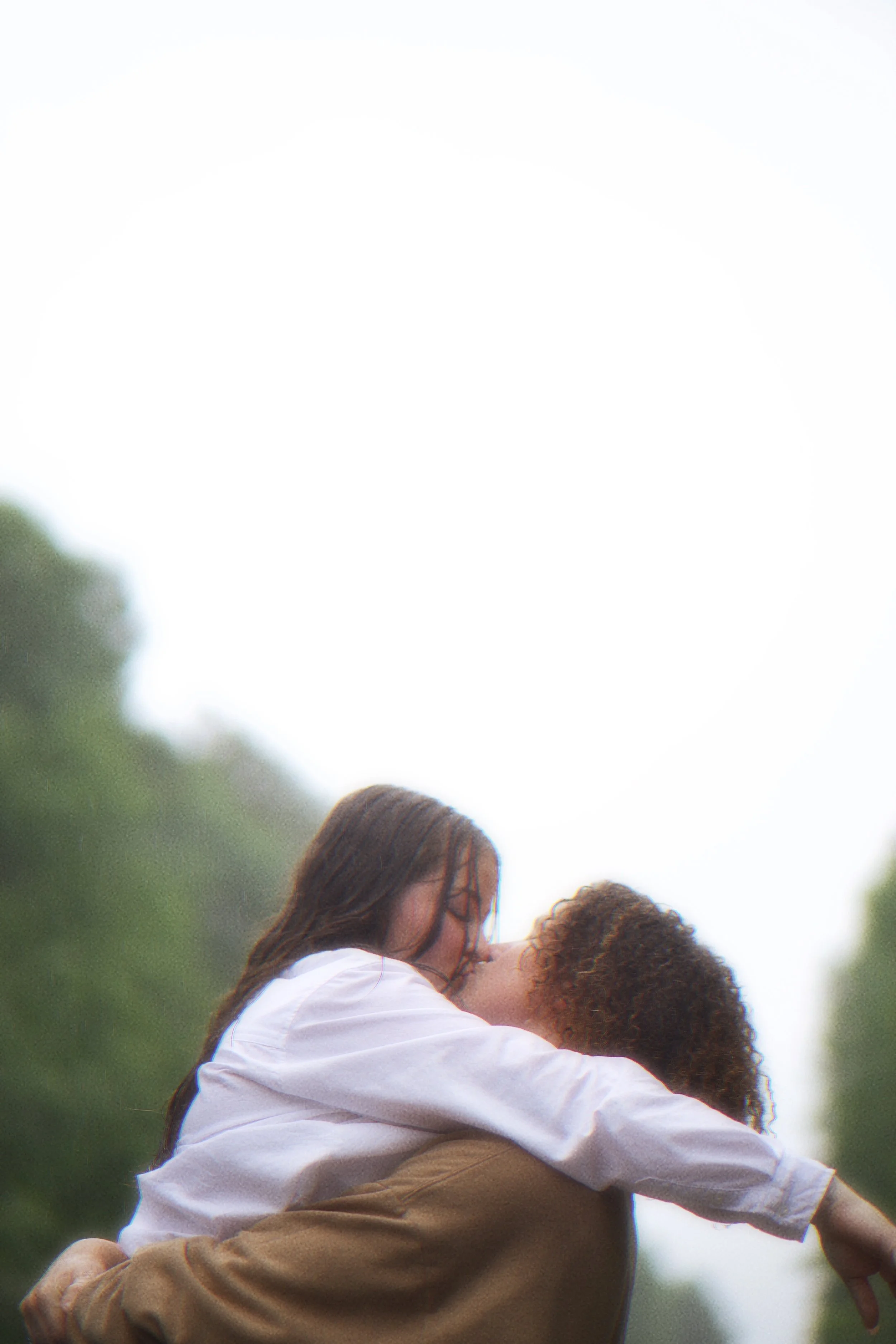 Two women sharing a kiss outdoors, one lifting the other, with blurry trees in the background.