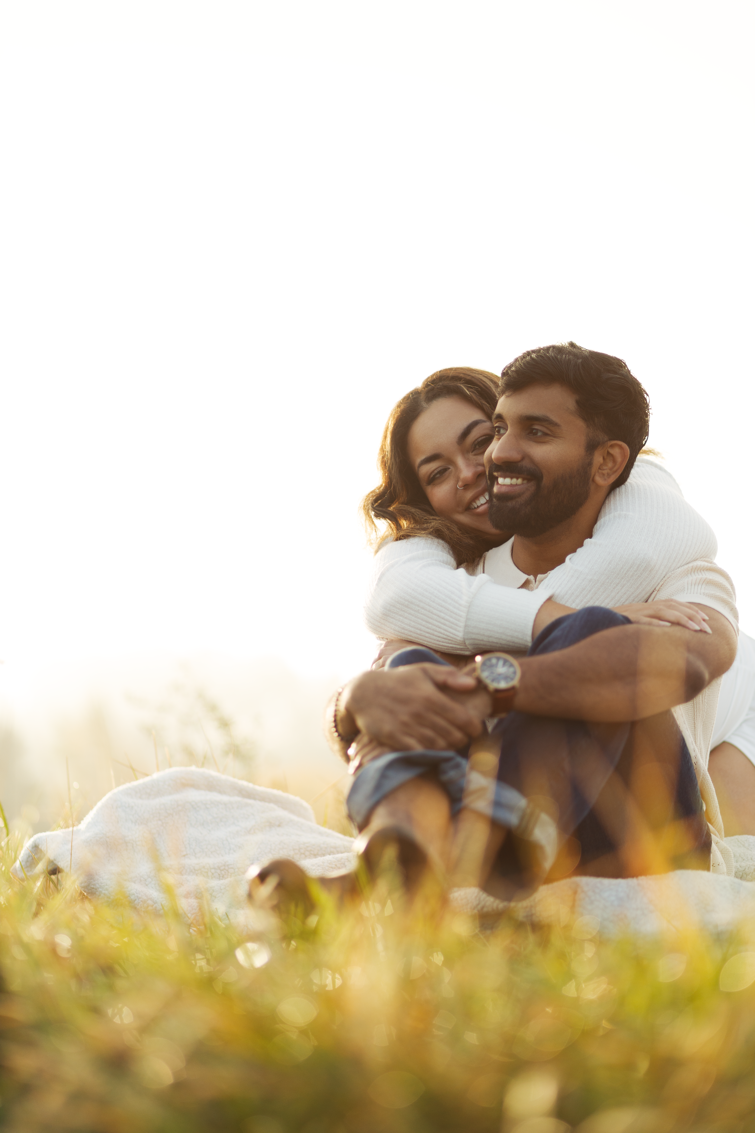 A happy couple sitting on the grass outdoors, embracing and smiling in the sunlight.