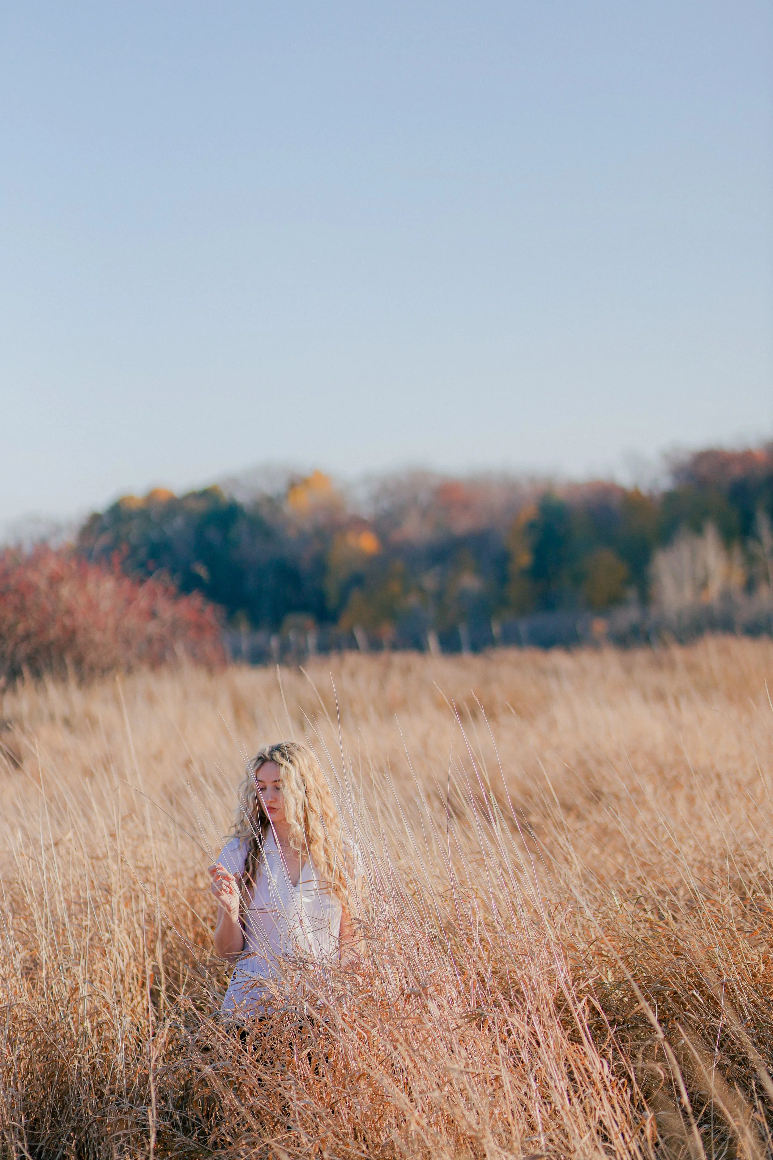 A woman with long blonde curly hair standing in a field of tall, dry grass on a clear day. She is wearing a white top and looking at something in her hand.