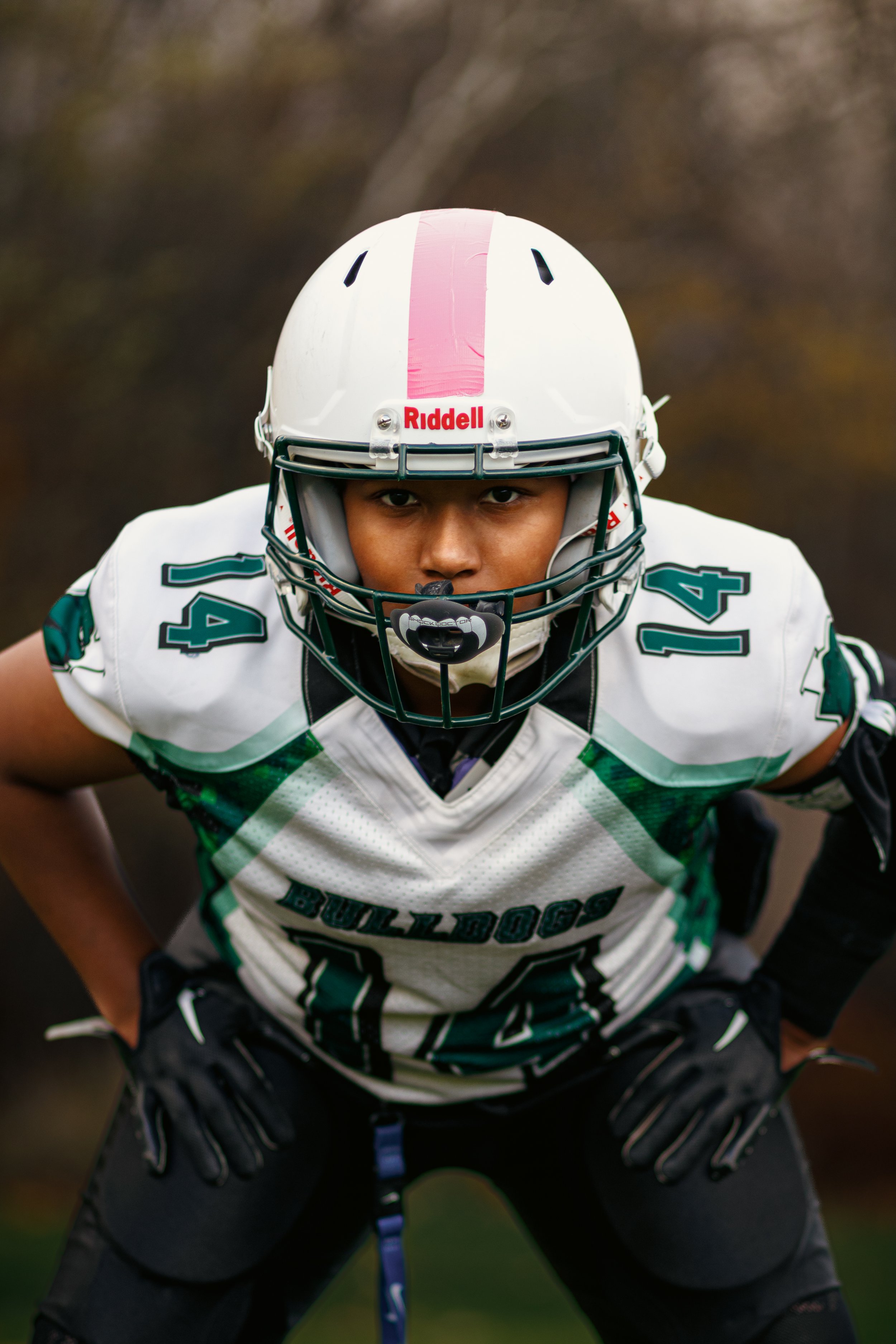 A football player wearing a white jersey with green and black accents, number 14, in a crouched stance, wearing a white helmet with a pink stripe and a face mask, on a football field with a blurred background.