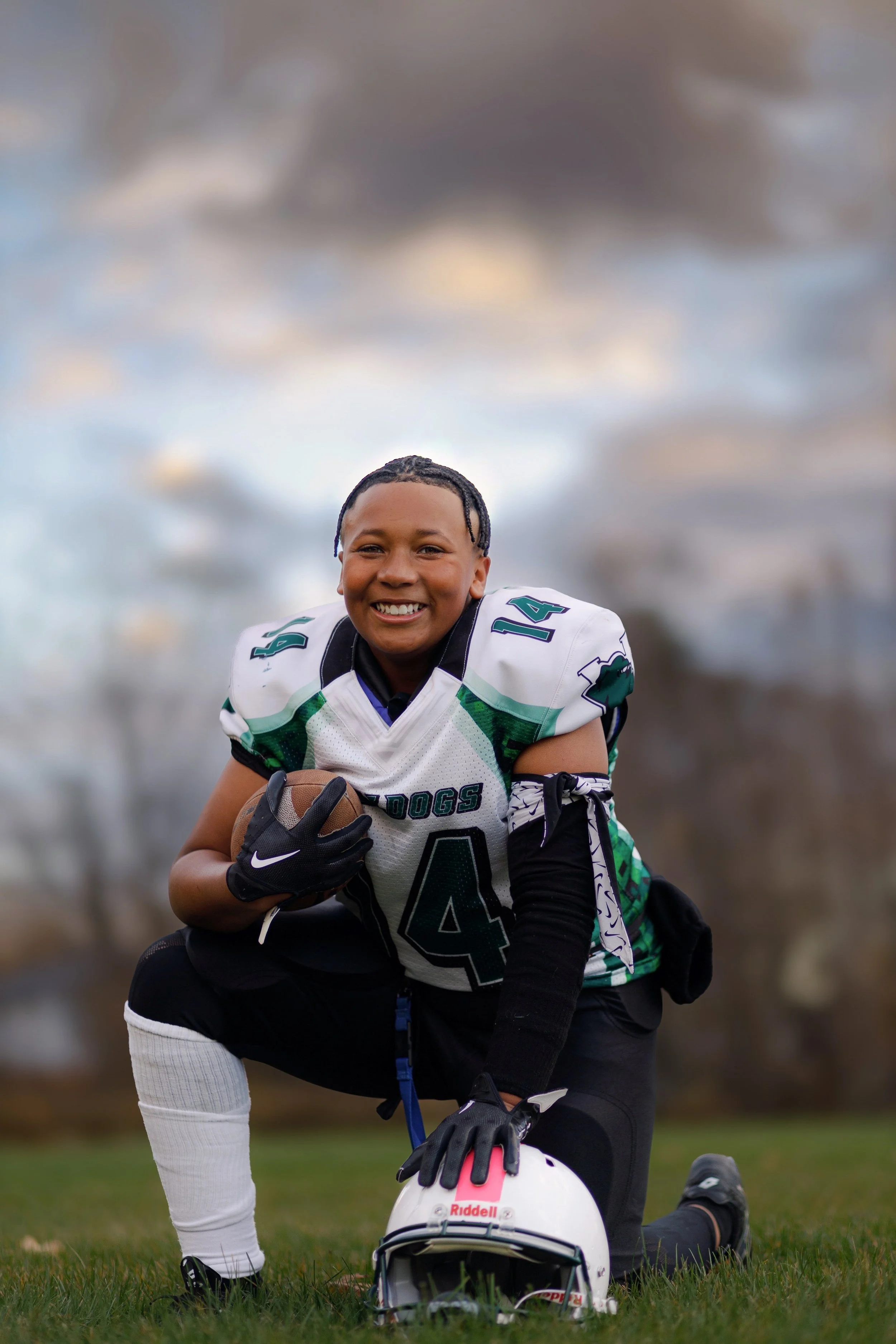 Young football player kneeling on grass field, smiling, holding a football in his right hand, wearing a white, green, and black jersey with the number 14, black gloves, black pants, and kneepads, with a white helmet placed on the grass in front of hi