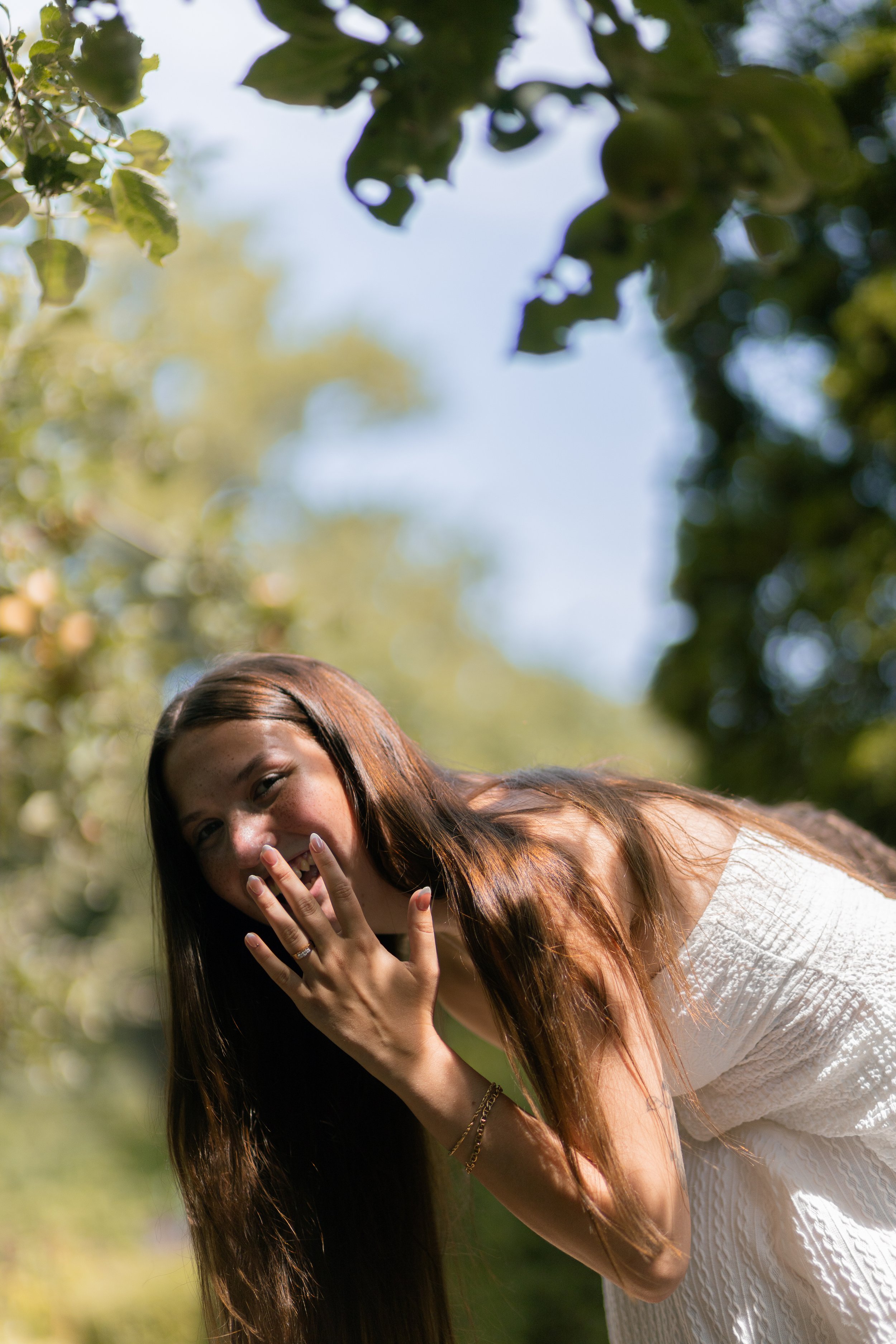 A young woman with long brown hair wearing a white dress, smiling and covering her mouth with her hand, standing outdoors among green trees under a blue sky.