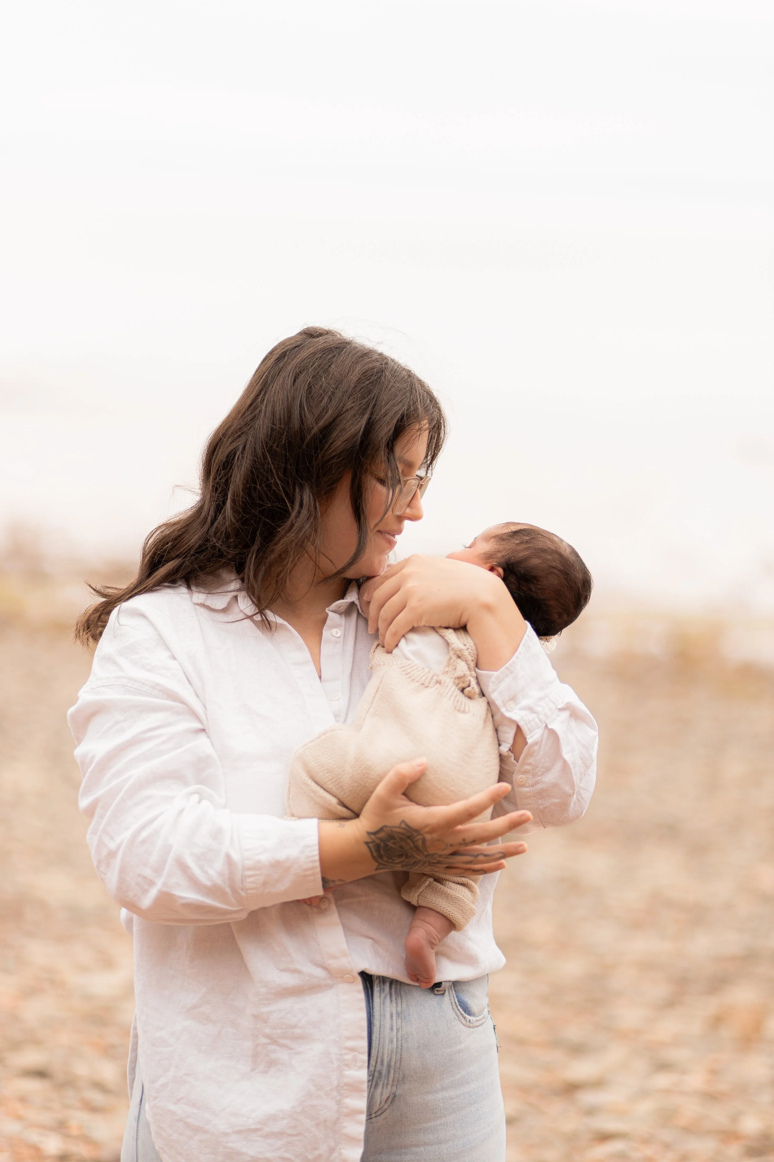 A woman with glasses and a tattoo on her hand holding a baby and looking at the baby in an outdoor setting.