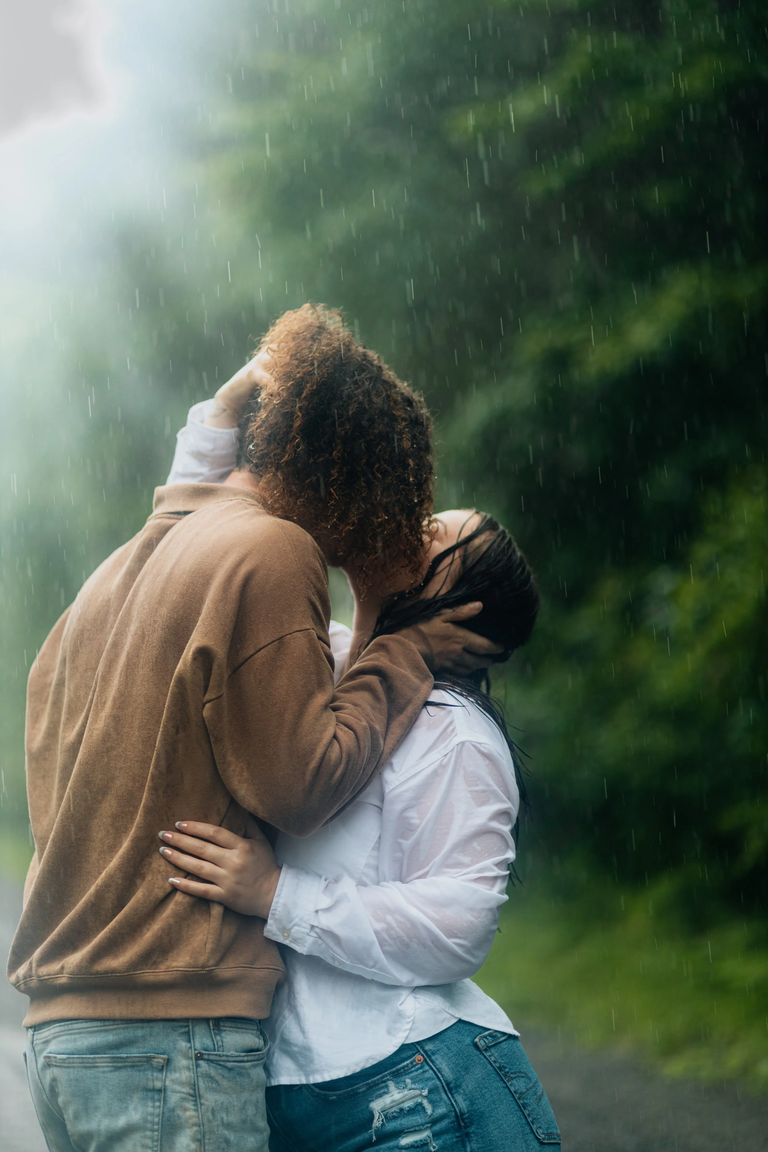 A couple kissing in the rain outdoors, with rain falling and greenery in the background.