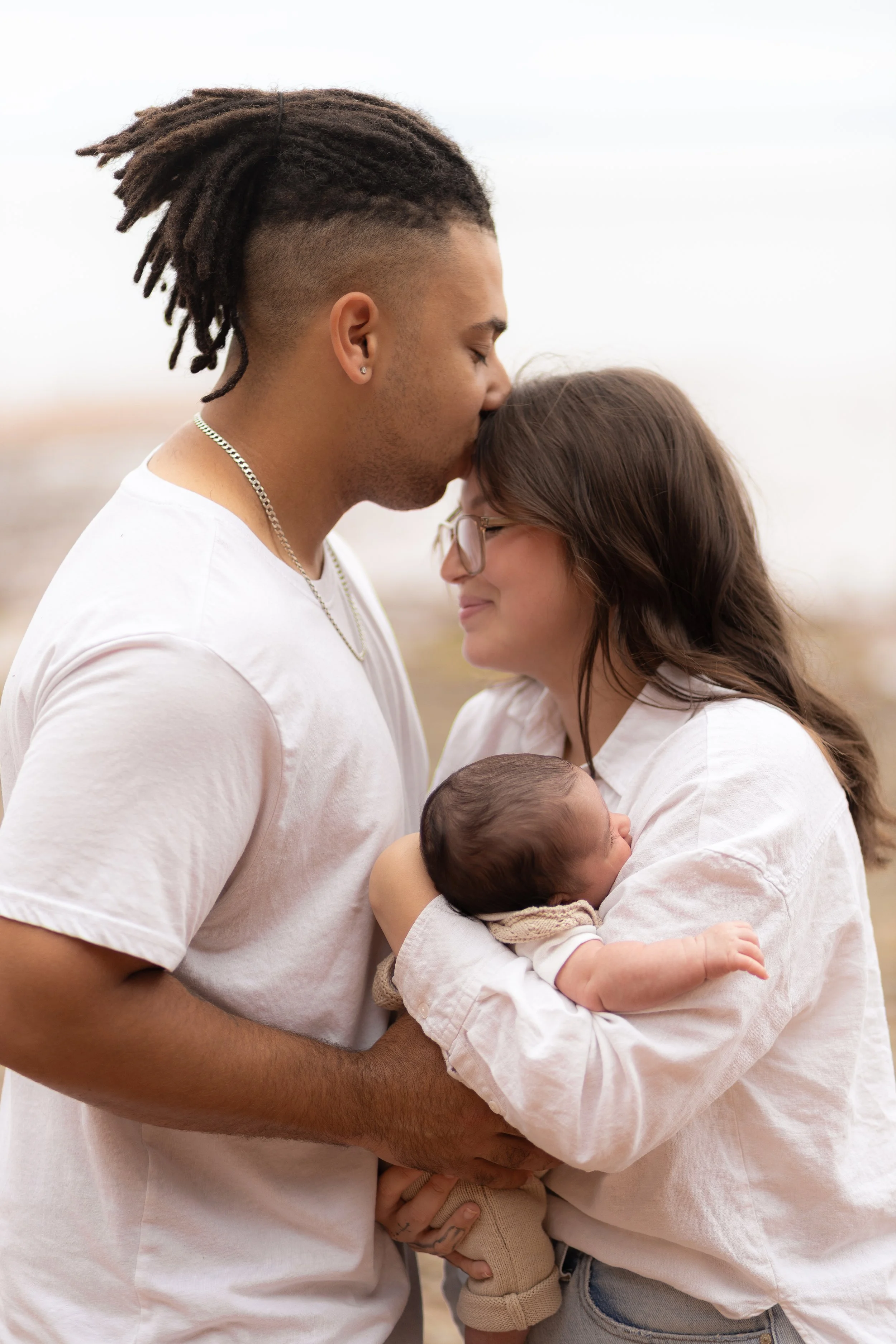 A couple holds their newborn baby close outdoors, the man kissing the woman's forehead as she smiles with her eyes closed, in a tender moment.