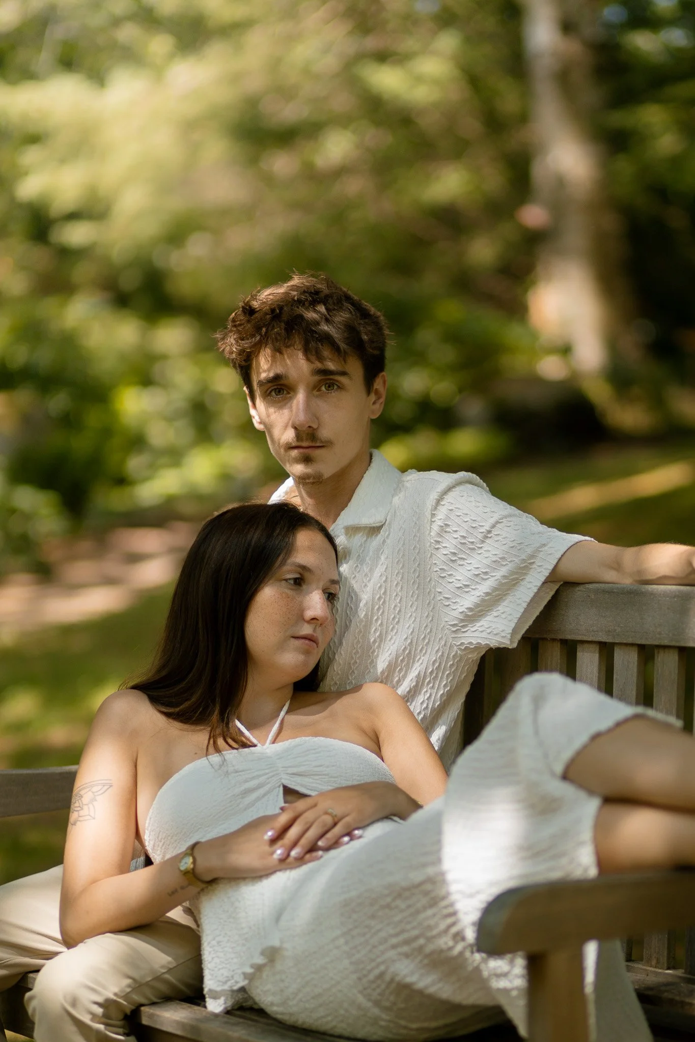 Young man and woman sitting on a park bench outdoors, the woman resting her head on the man's shoulder, both wearing white clothing, surrounded by green trees.