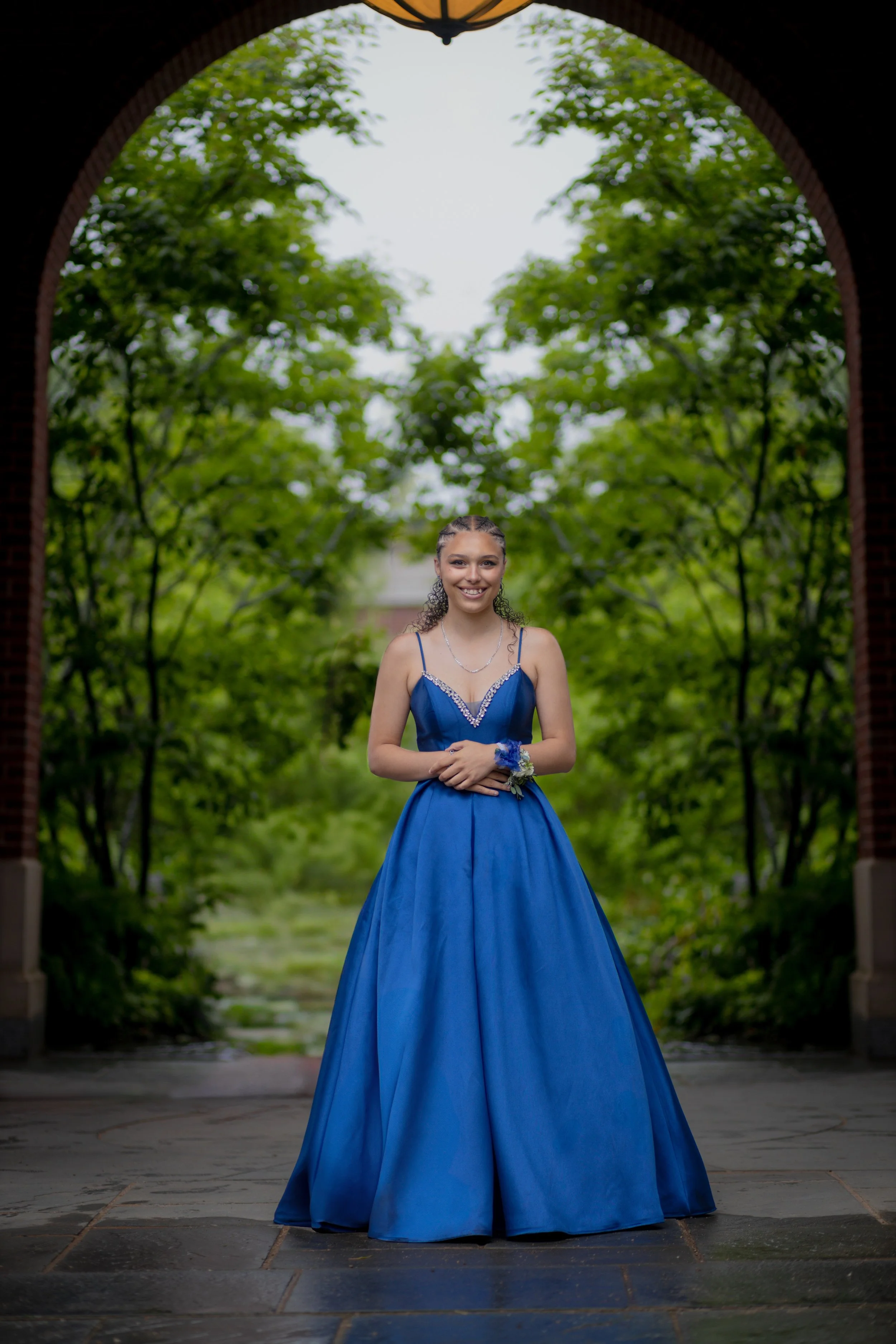 A young woman in a blue formal gown standing under an archway surrounded by green trees. She is smiling, wearing a corsage on her wrist, and jewelry, with her hands clasped in front of her.