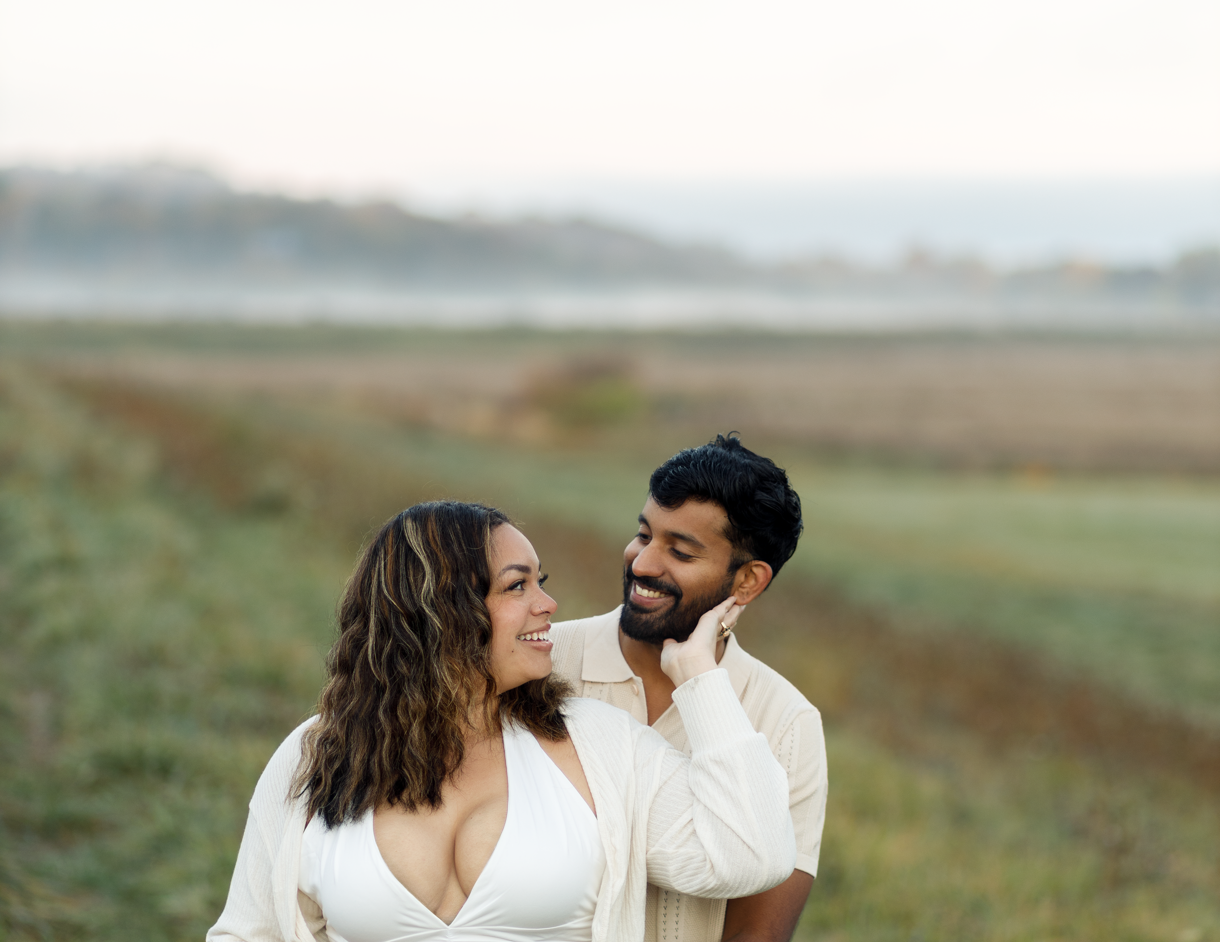 A smiling couple standing outdoors in a field, with the man touching the woman's face while they look at each other, with a scenic landscape in the background.