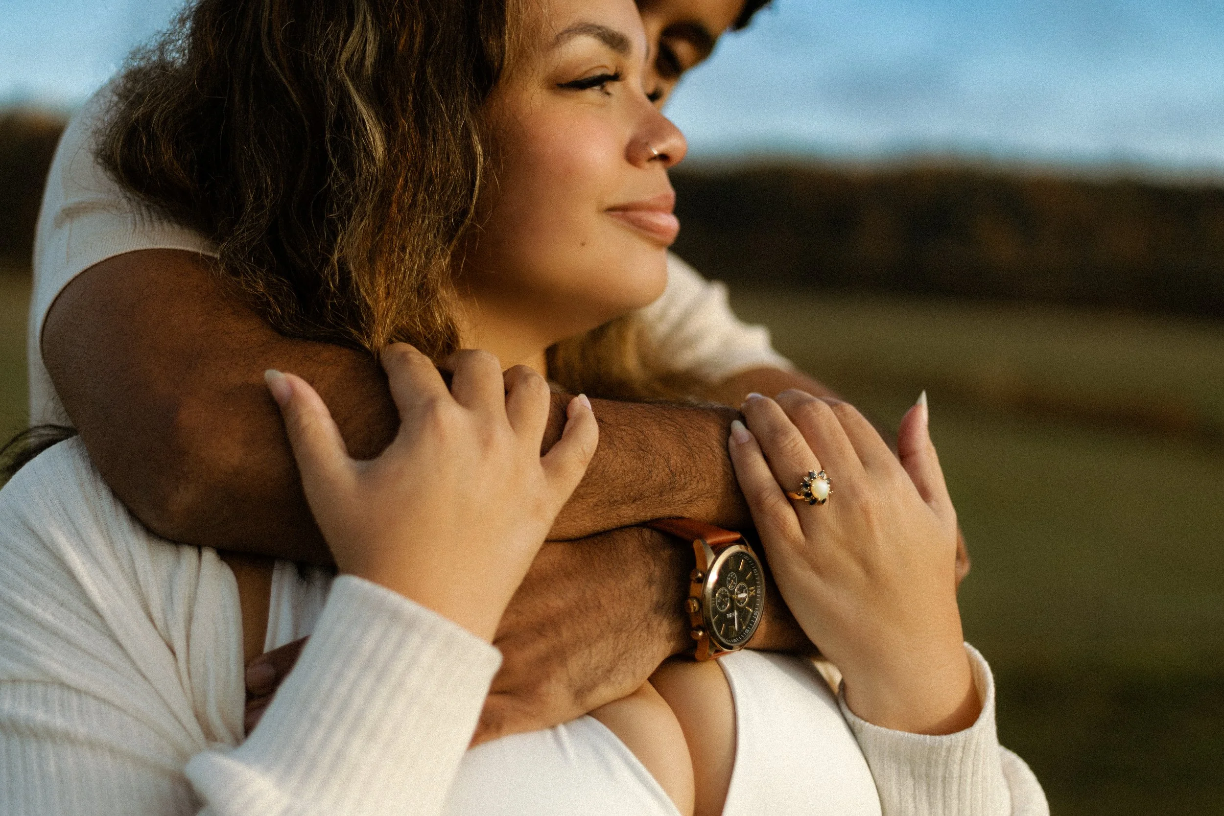 A woman with curly hair and a nose ring embraces a man from behind, with her hands on his arms, standing outdoors during sunset.