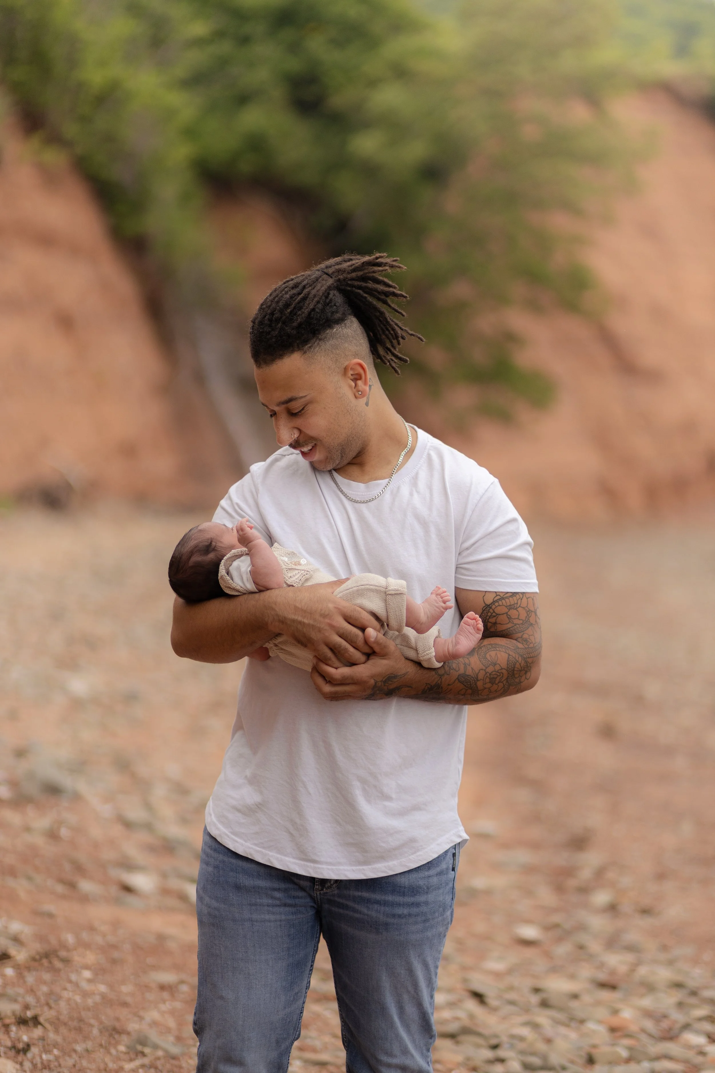 A young man with dreadlocks and tattoos, smiling and holding a newborn baby outdoors on a dirt path with green trees and a brown hillside in the background.