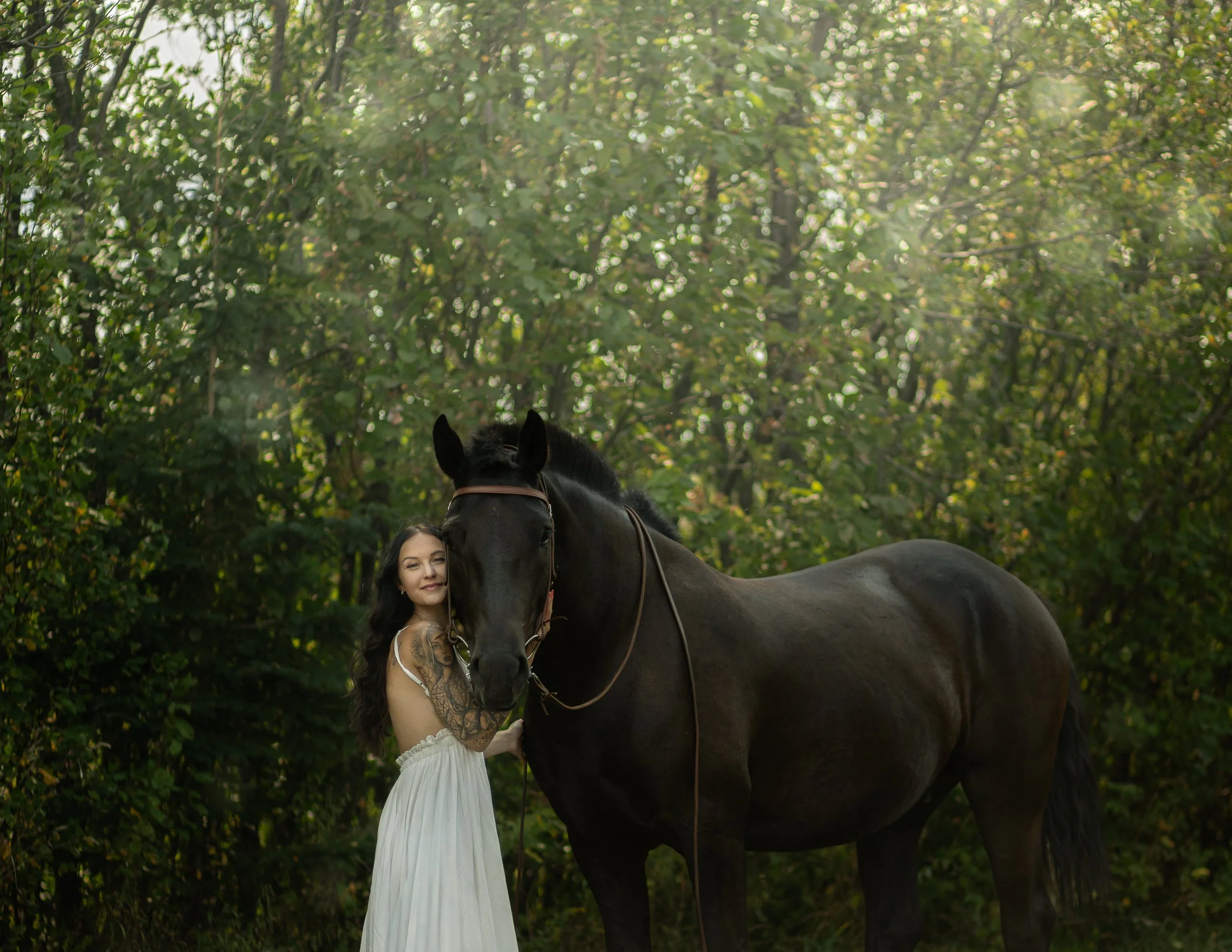 A woman with long dark hair, wearing a white dress, standing next to a large black horse in a green wooded area.
