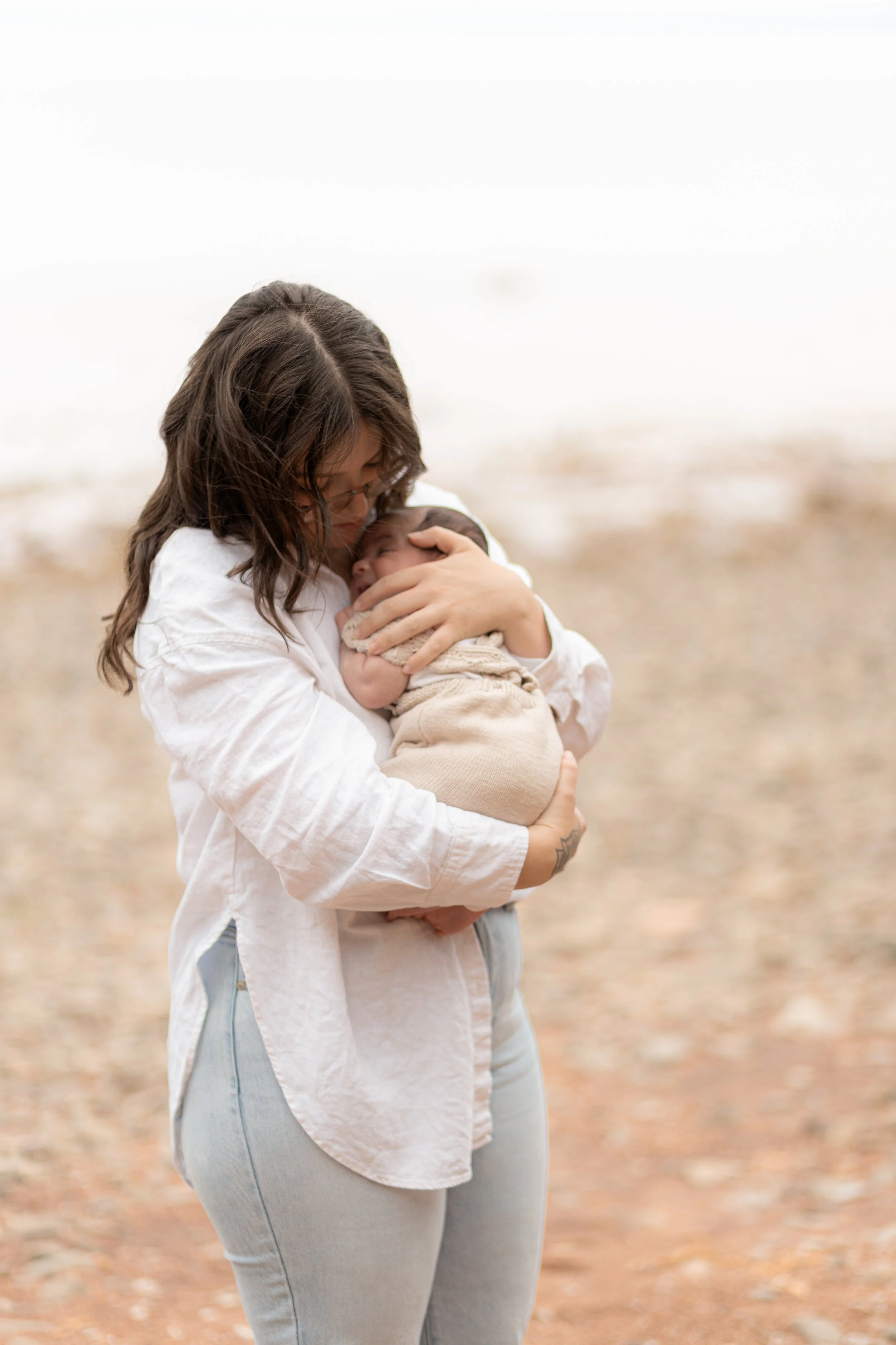 A woman holding a newborn baby on a beach, with the shoreline in the background.