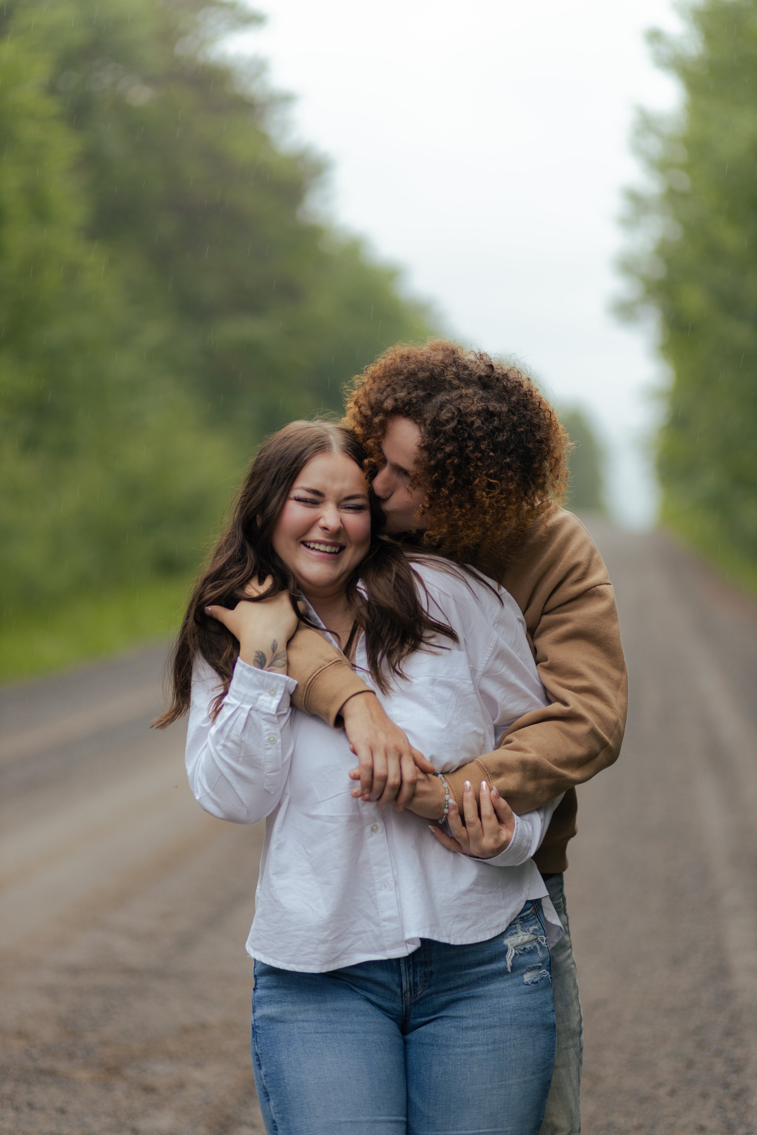 A couple sharing a joyful moment on a rainy day on a dirt road surrounded by green trees, with the man kissing the woman's cheek.