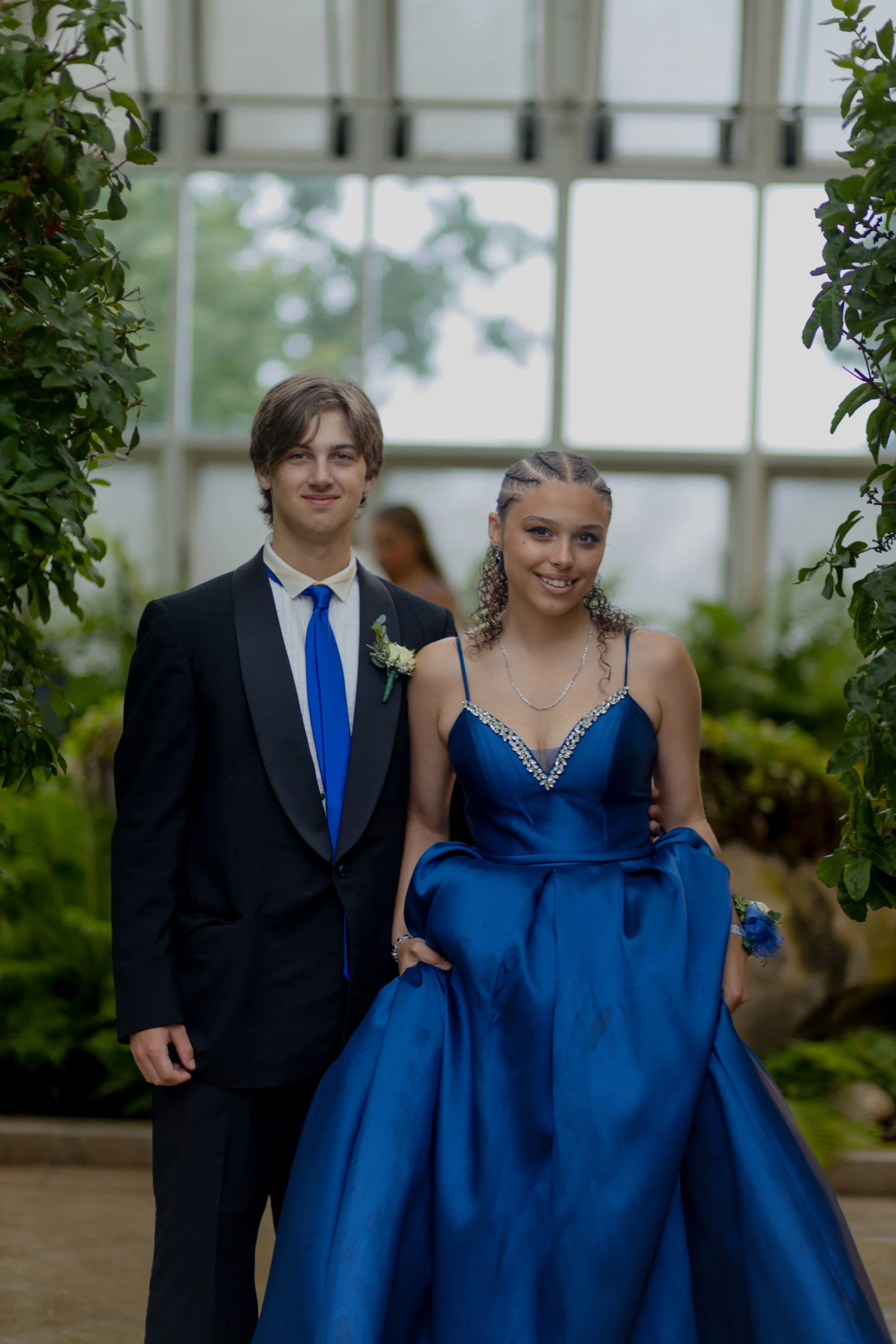 Young couple dressed in formal attire at a greenhouse or conservatory, with greenery and large windows in the background.