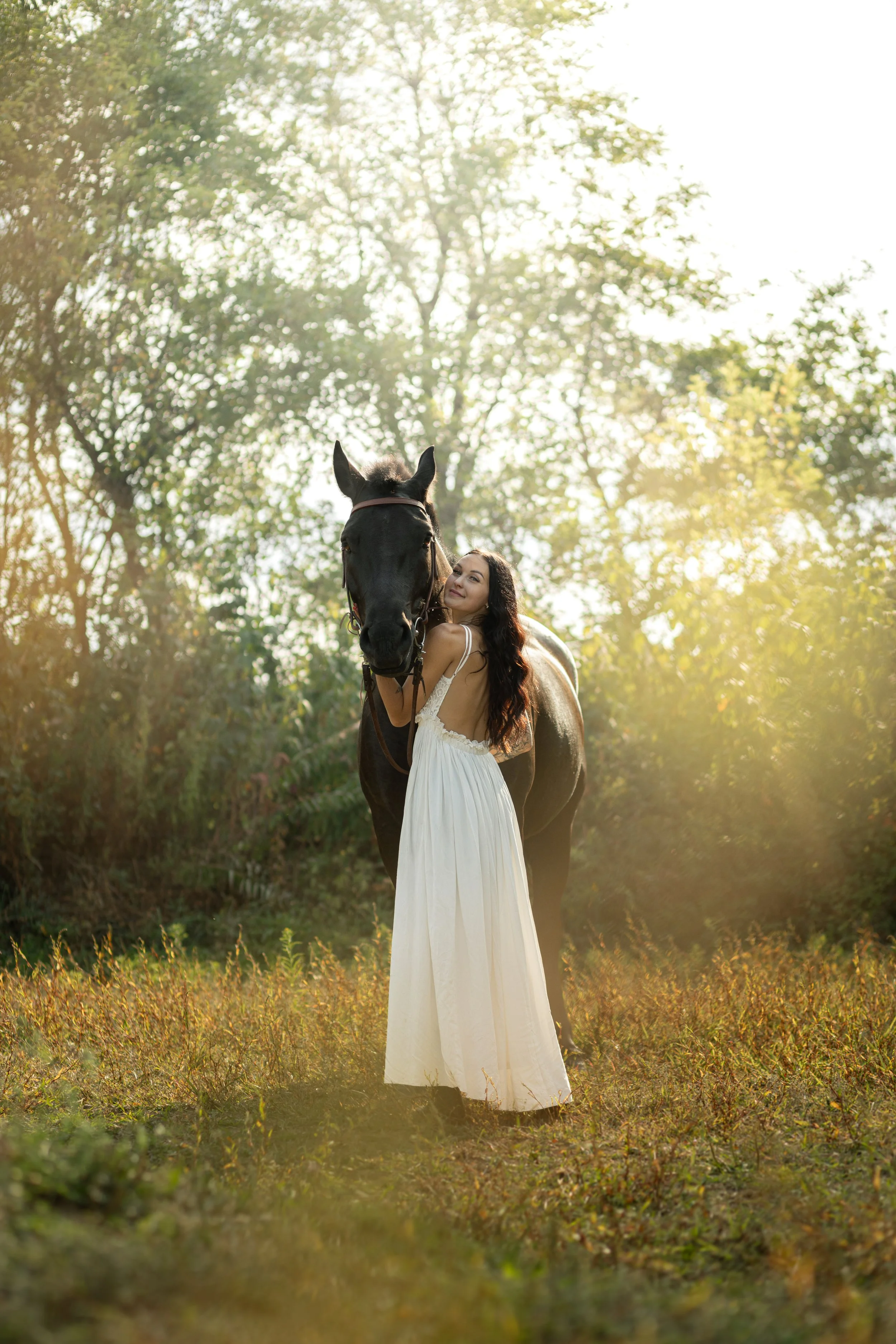 A woman in a white dress hugging a black horse outdoors with sunlight filtering through trees in the background.