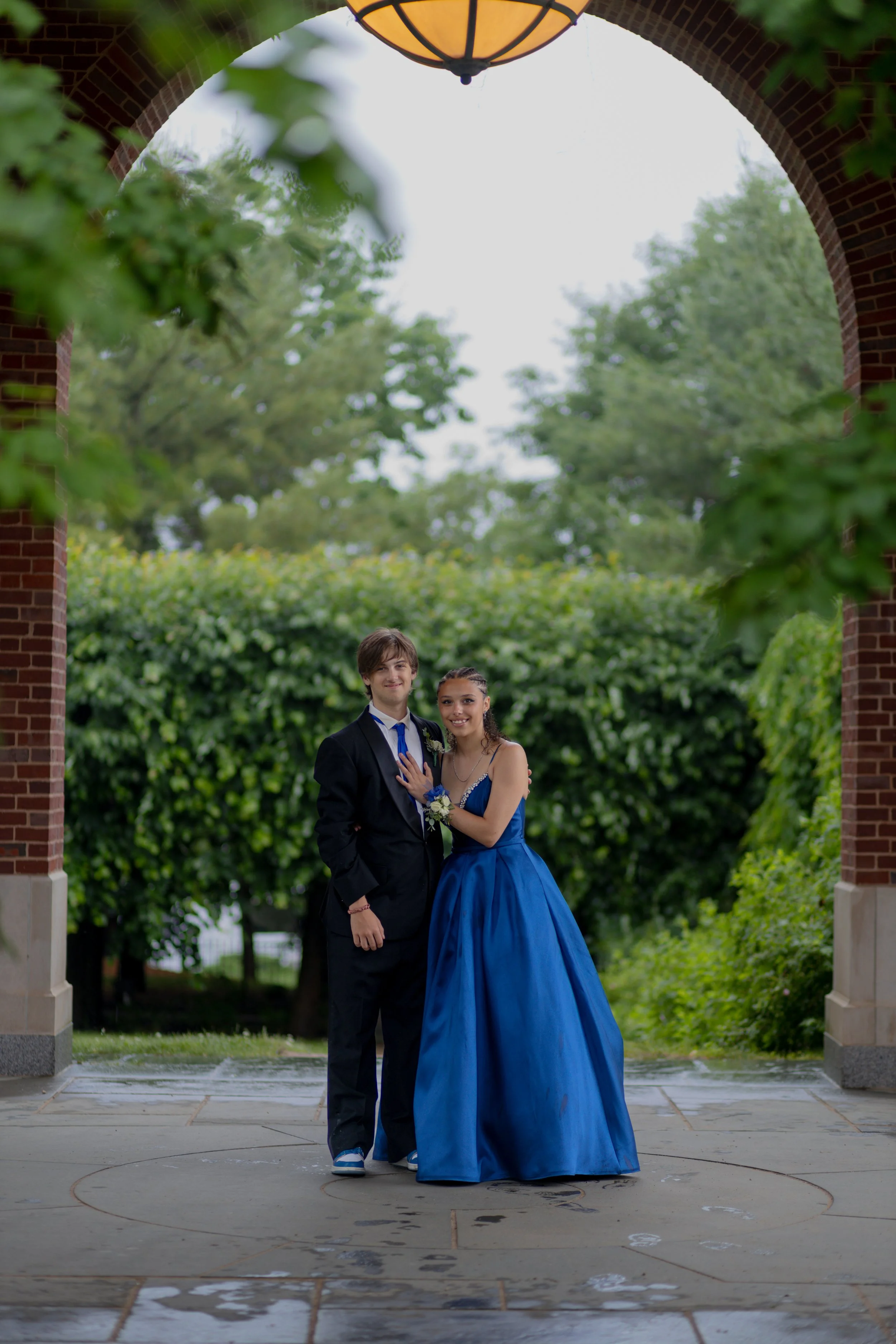 Young couple in formal attire standing under a brick archway, with green trees and foliage in the background, on a damp pavement.
