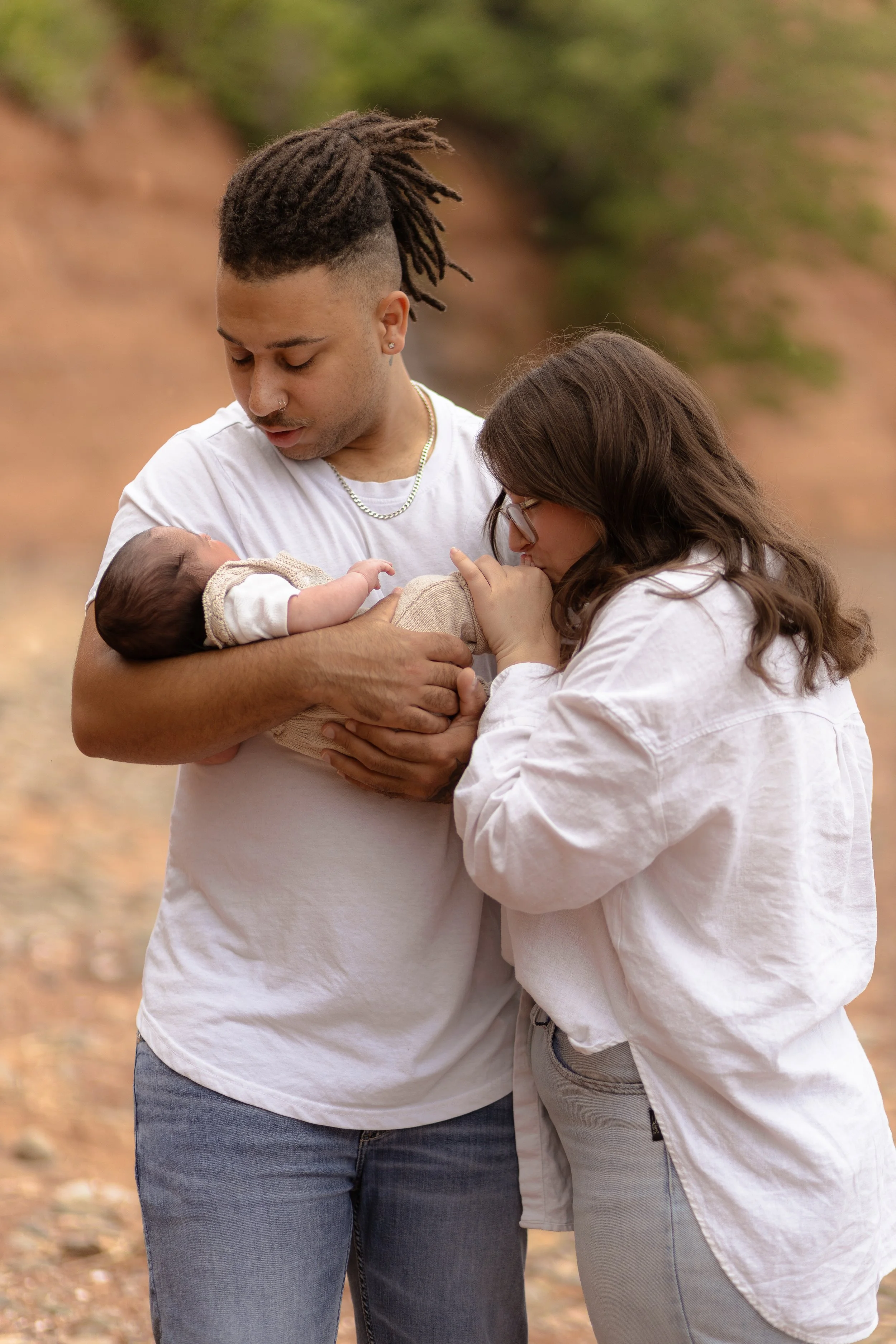 A young couple holding a newborn baby outdoors on a fall day, with trees and leaves in the background.