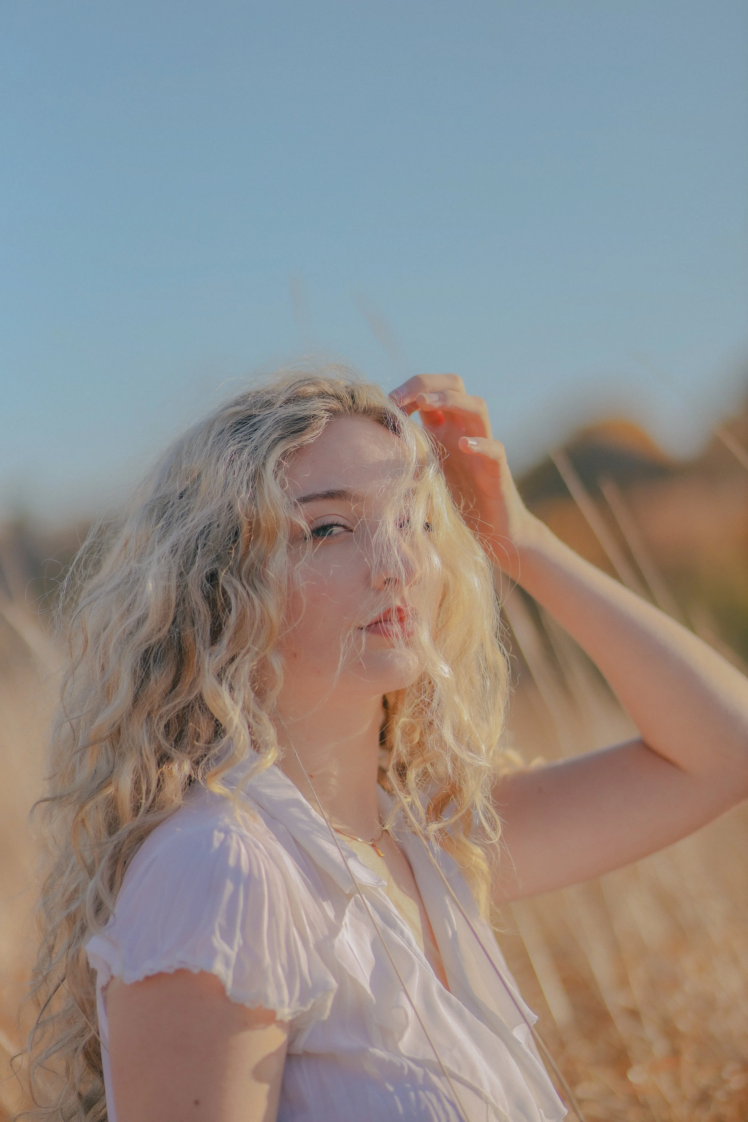 A young woman with blonde curly hair standing in a field under a clear blue sky, wearing a white blouse, with her hand touching her hair.