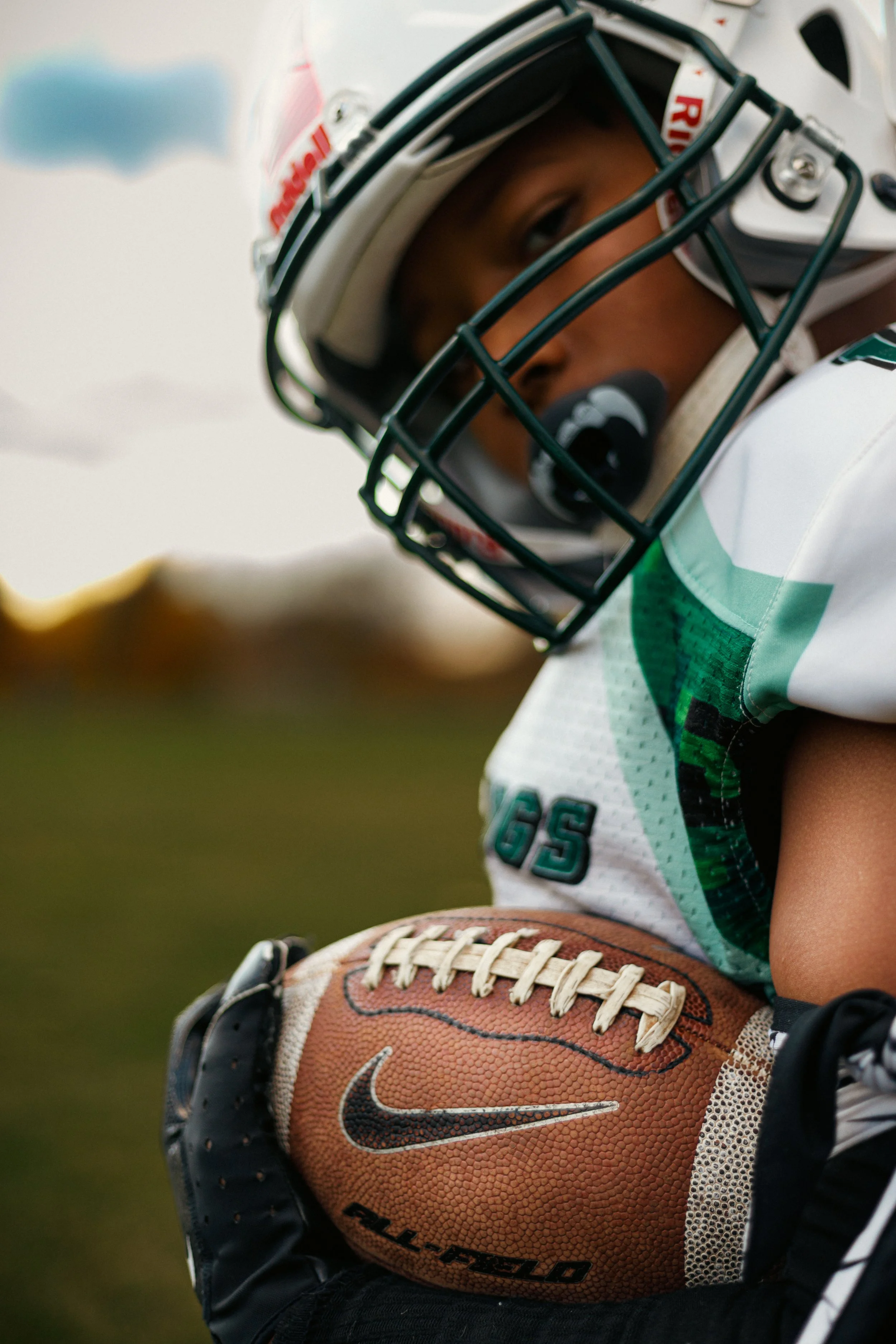 Close-up of a young football player wearing a helmet and jersey, holding a Nike football with a sunset-lit sky in the background.