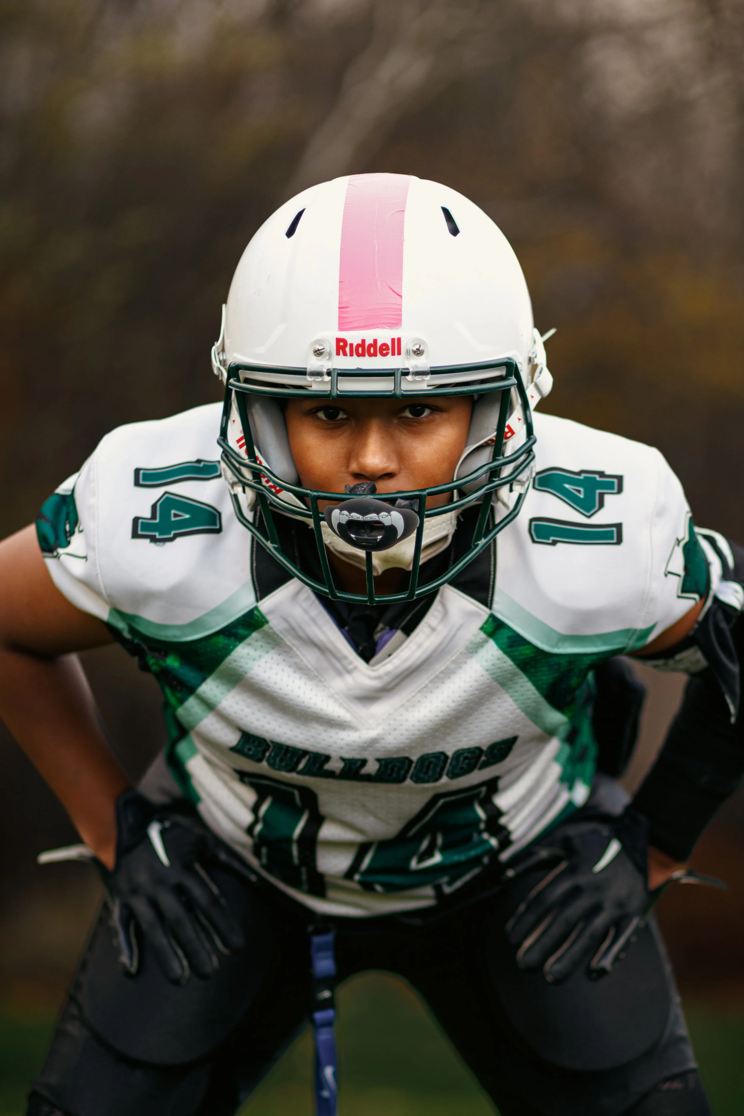 A football player in a white and green jersey number 14, wearing a white helmet with a pink stripe, in an athletic stance outdoors.