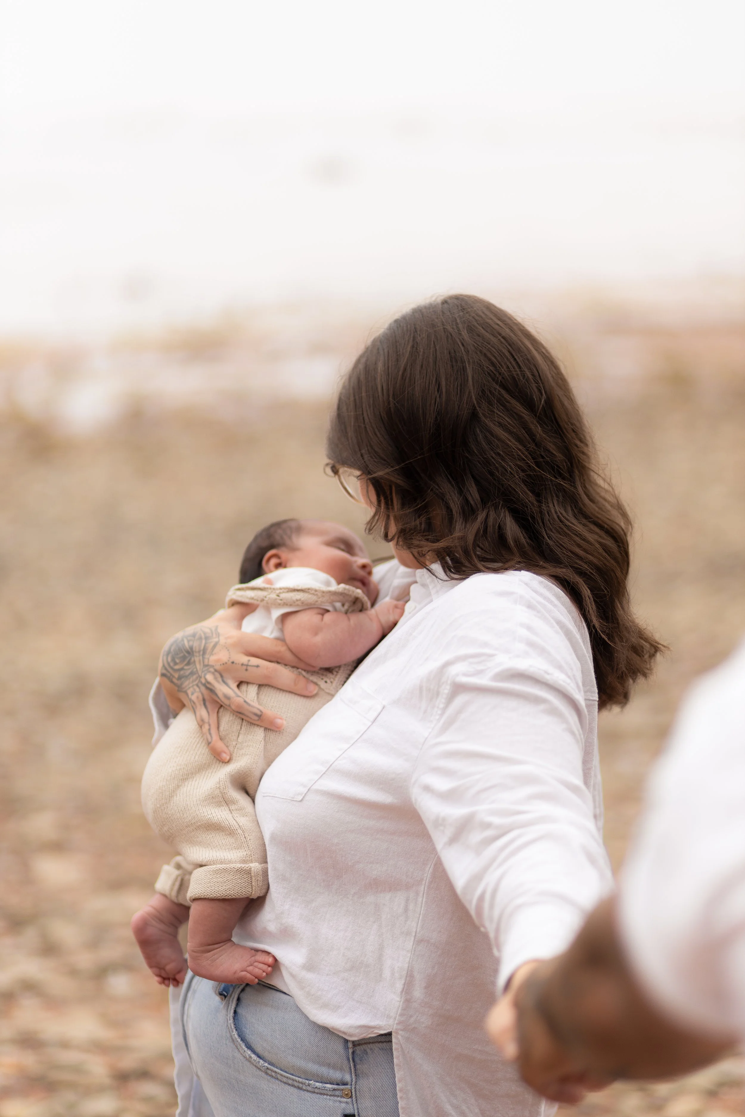 A woman with long dark hair and glasses holding a newborn baby outside on a cloudy day.