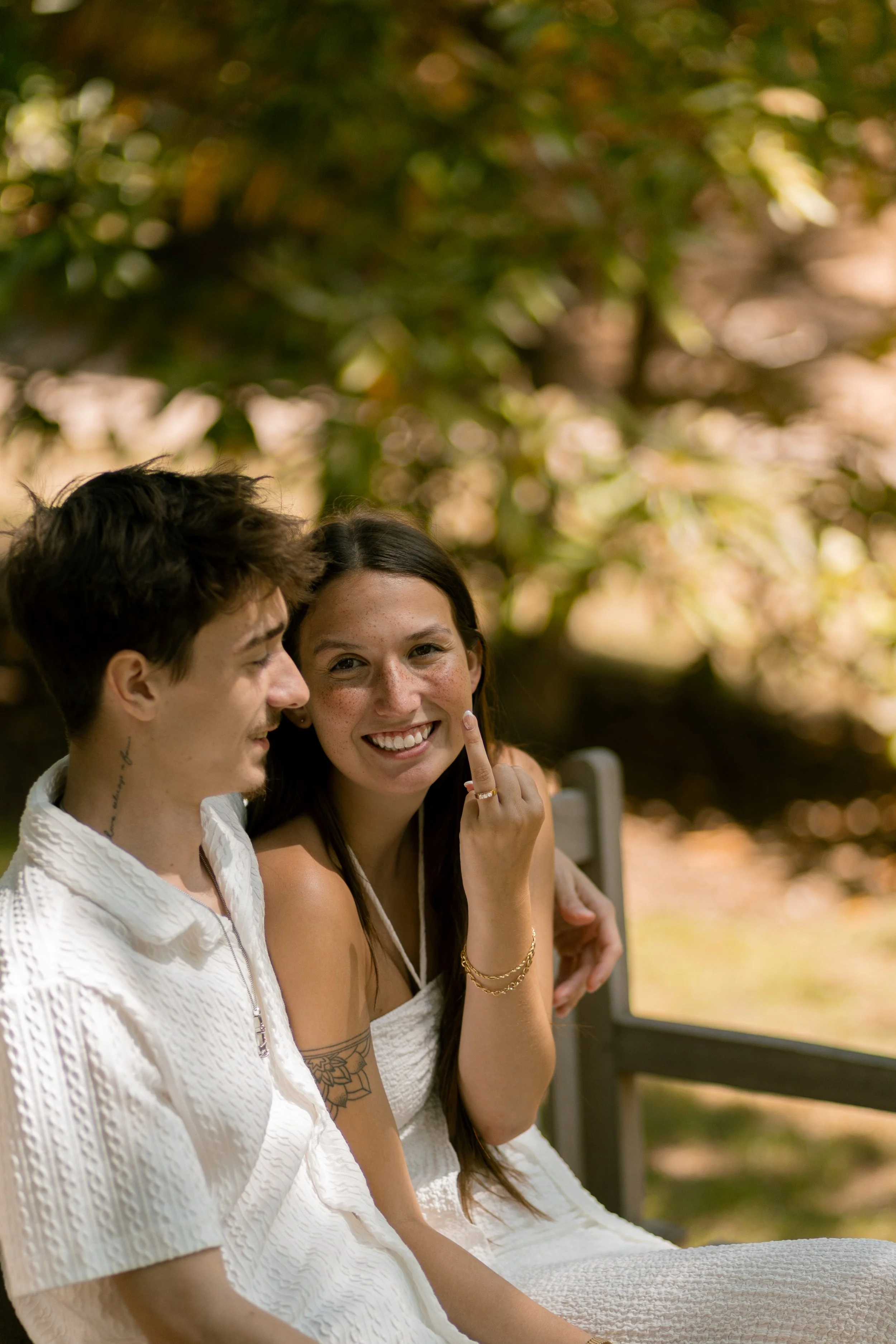 A young woman with long dark hair smiling and showing her middle finger while sitting outdoors next to a young man with short dark hair. The woman wears gold jewelry, tattoos, and a white dress, and they are on a bench with a blurred natural backgrou