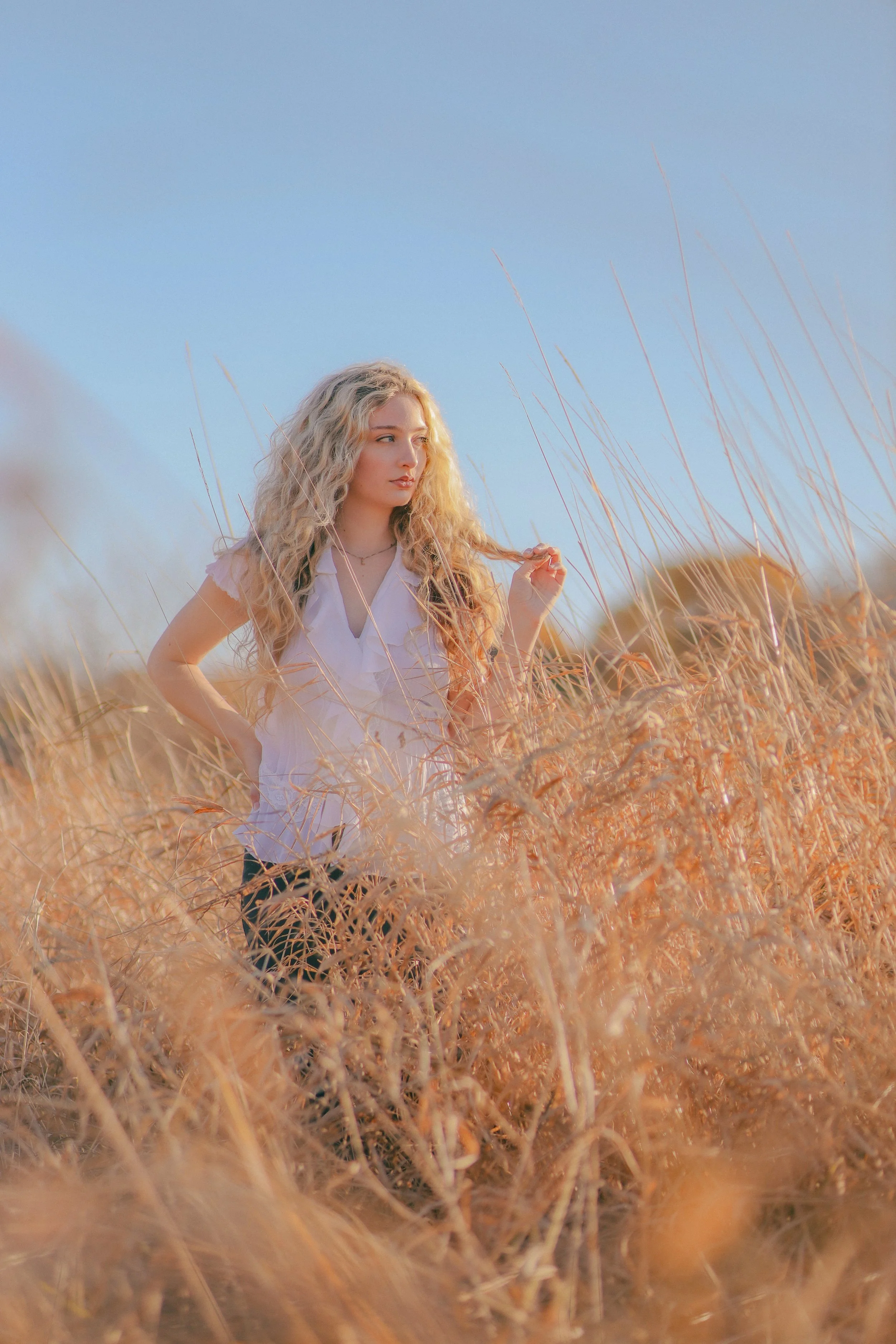 A young woman with long, curly blonde hair standing in a field of tall, dry grass under a clear blue sky.