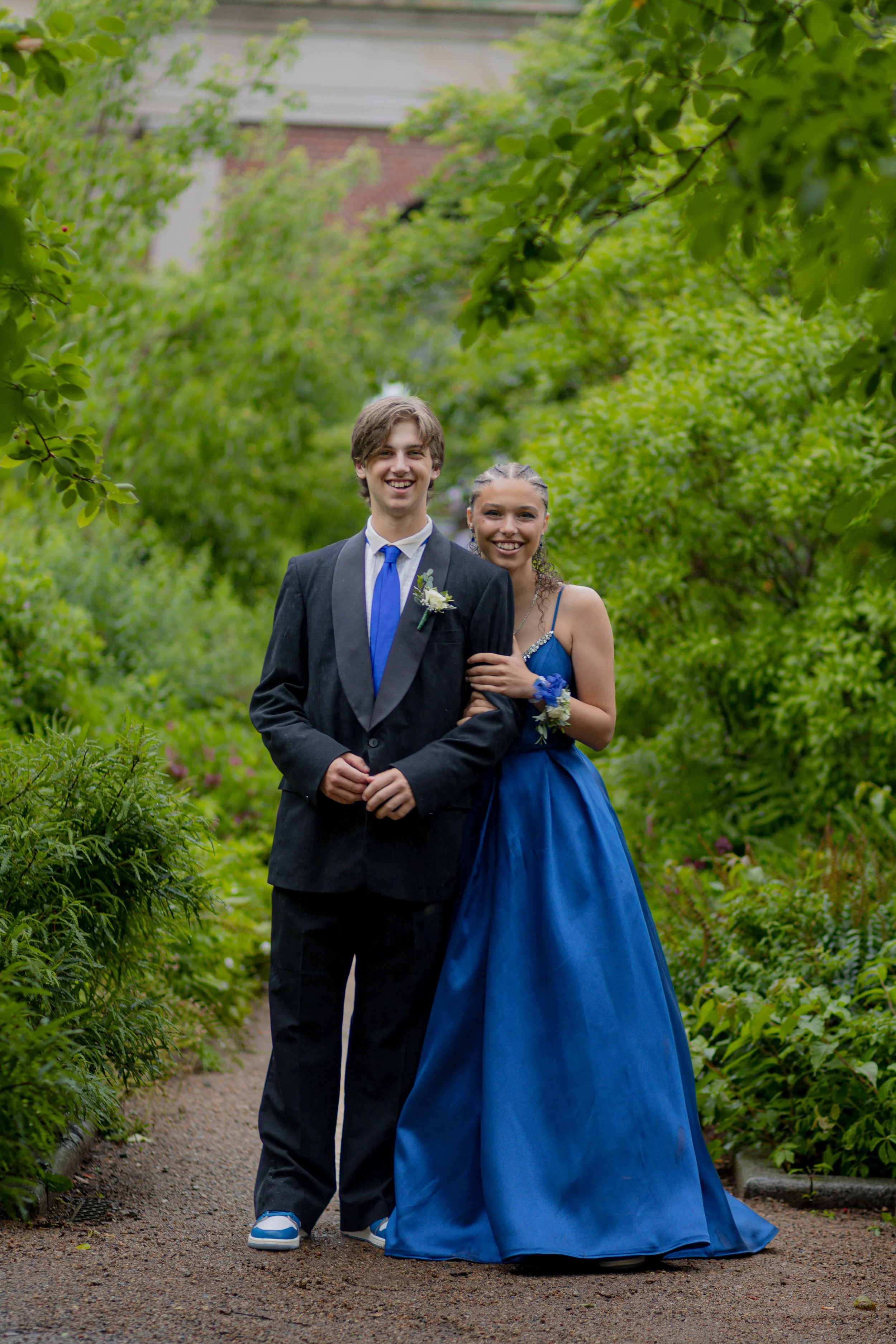 Two teenagers, a boy and a girl, dressed in formal attire, standing on a garden path surrounded by lush green foliage. The boy wears a dark suit with a blue tie, and the girl wears a long blue dress with a corsage. Both are smiling and looking at the