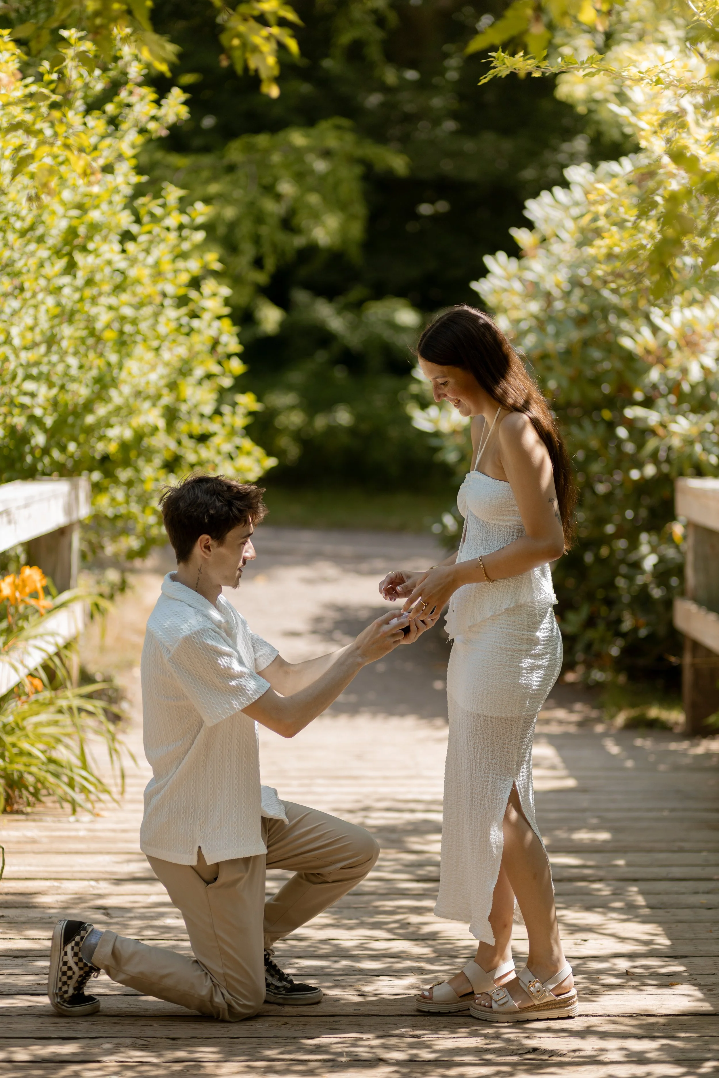 A man is proposing to a woman on a wooden pathway in a lush, green outdoor setting. The man is kneeling on one knee, holding the woman's hand as he proposes. The woman is smiling, looking down at him, wearing a white dress, and sandals. The scene is 