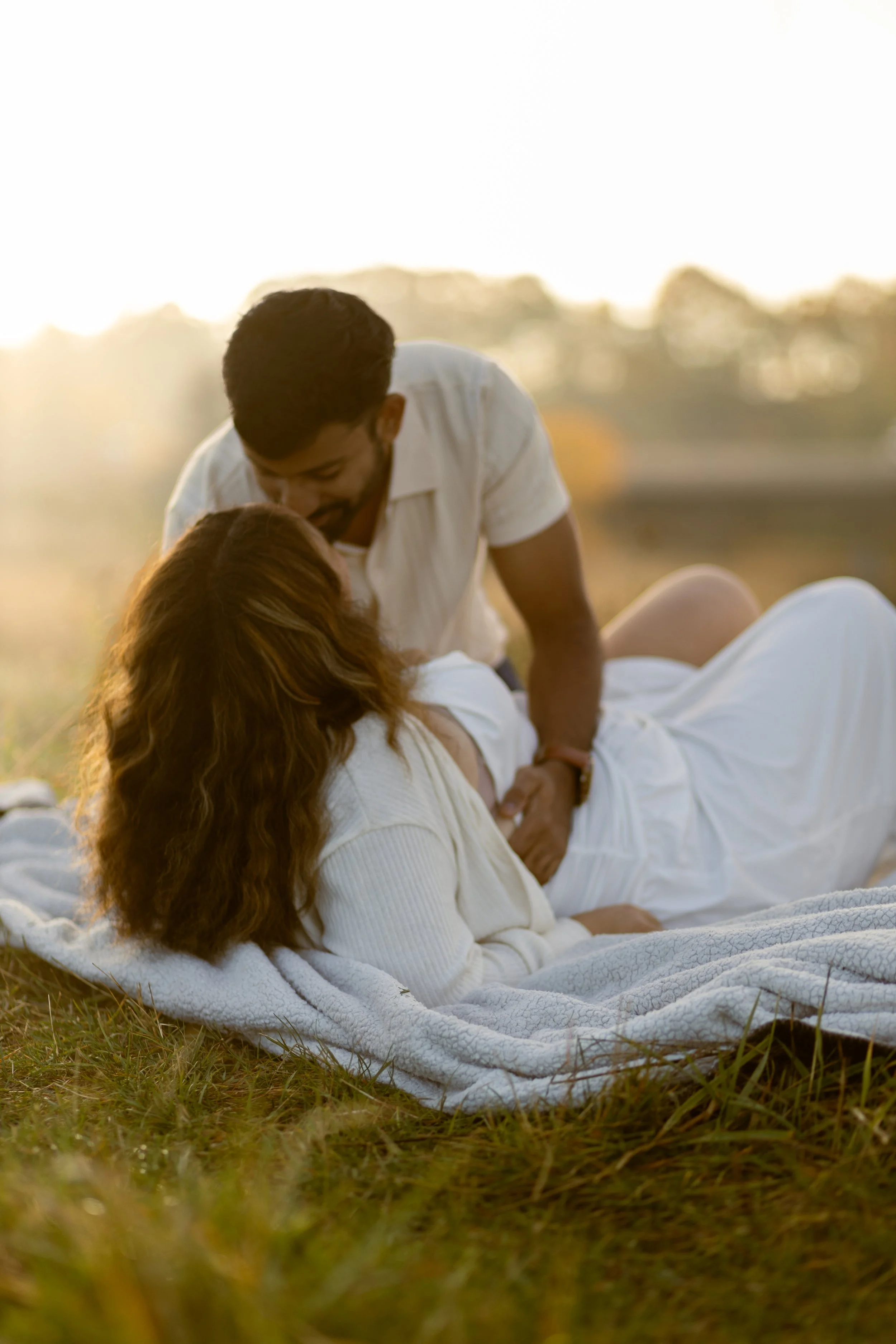 A couple is lying on a blanket outdoors during sunset, with the man leaning over the pregnant woman, in a tender moment.