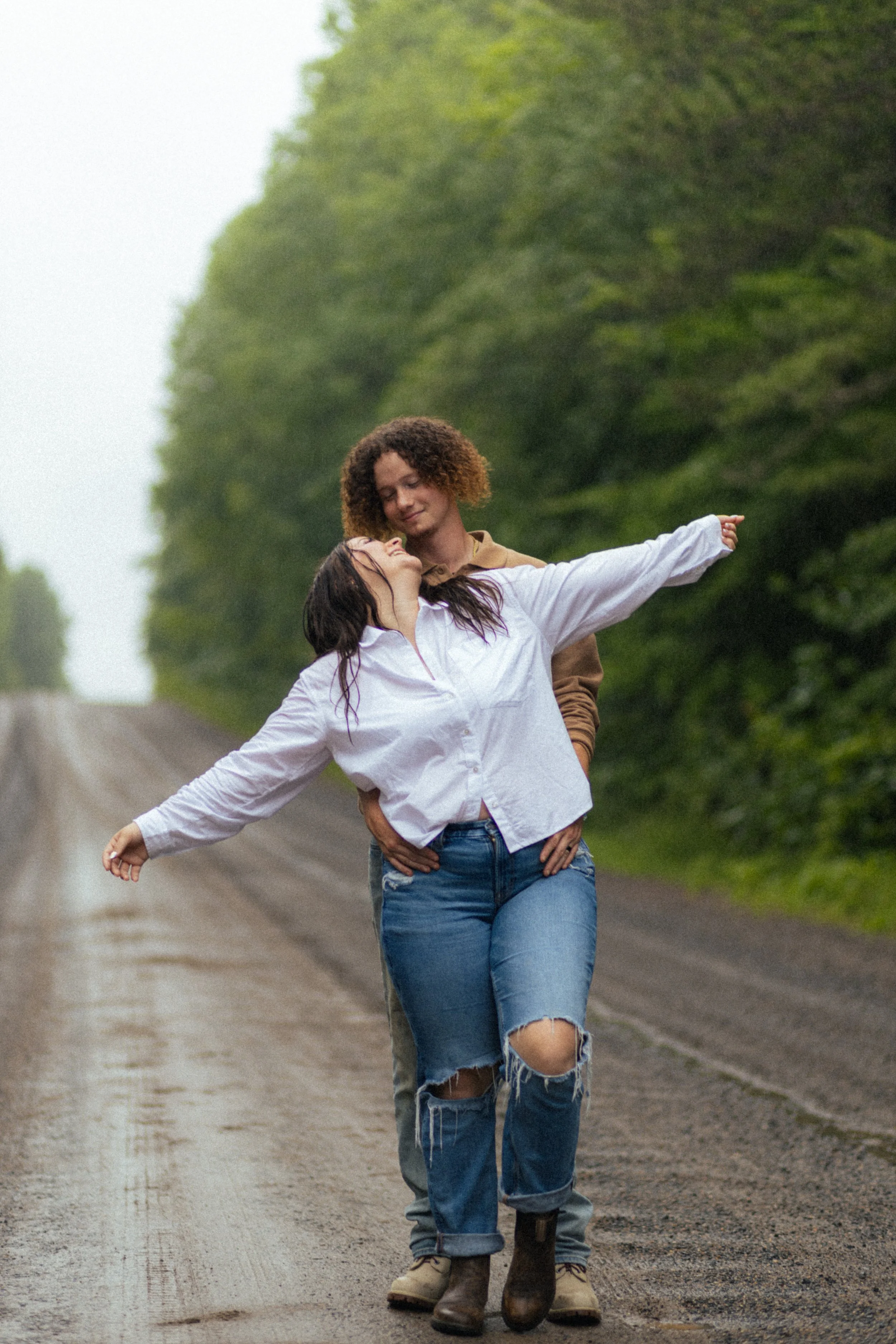 A couple standing on a dirt road surrounded by forest, engaging in a romantic embrace. The woman is leaning back with arms outstretched, wearing a white shirt and ripped jeans, while the man stands behind her with hands in pockets.