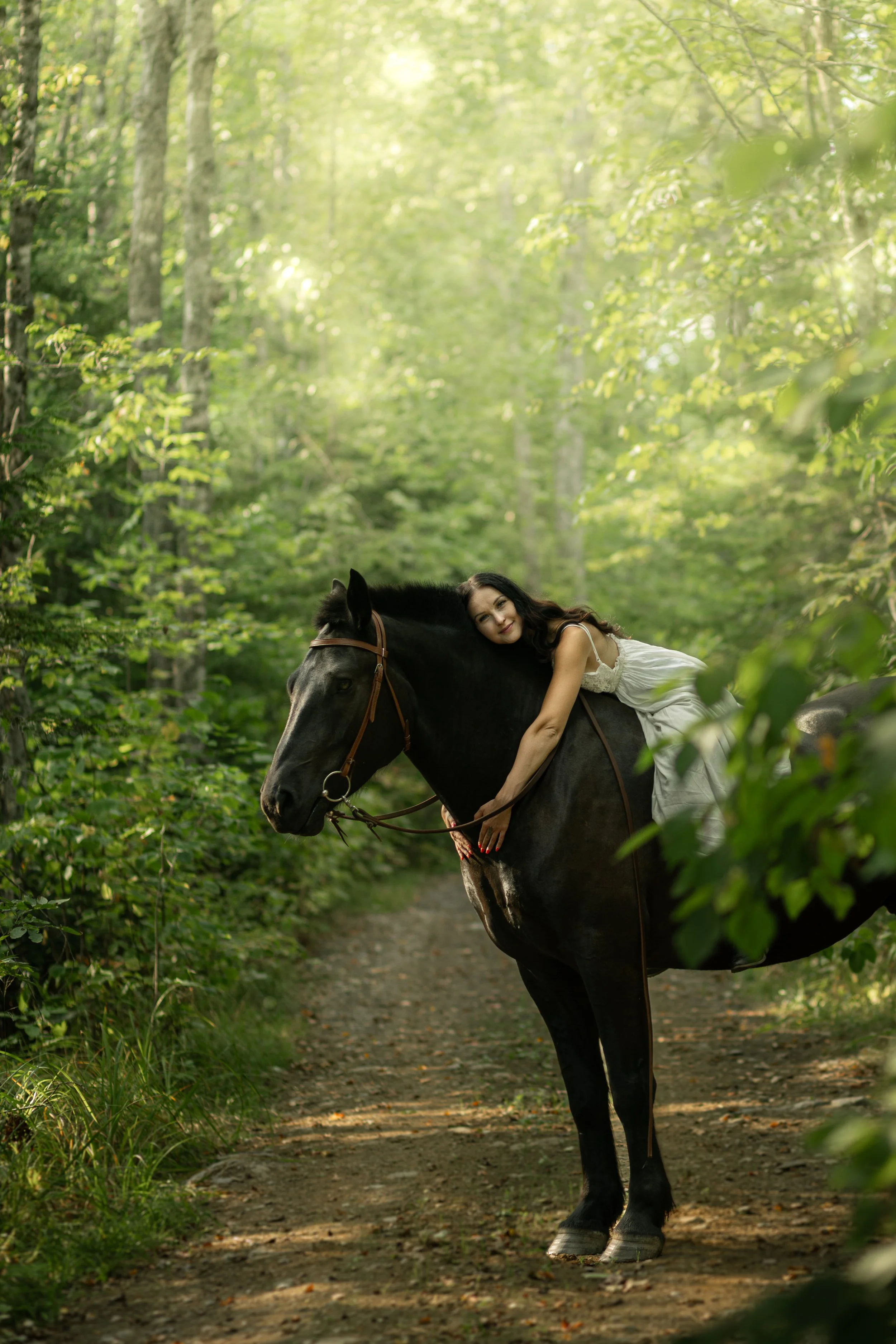 A woman in a white dress rests on a black horse on a dirt trail in a lush green forest.